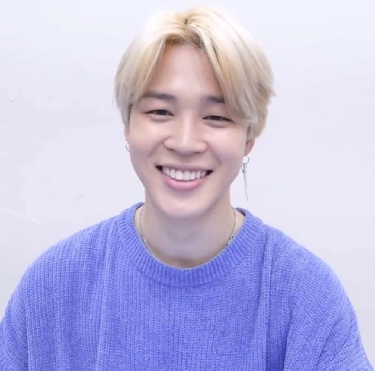 Jimin Aesthetic 桌布 壁紙 — 一位開朗的金髮年輕男子，面帶燦爛笑容，穿著質感豐富的紫色毛衣，背景為純白色。