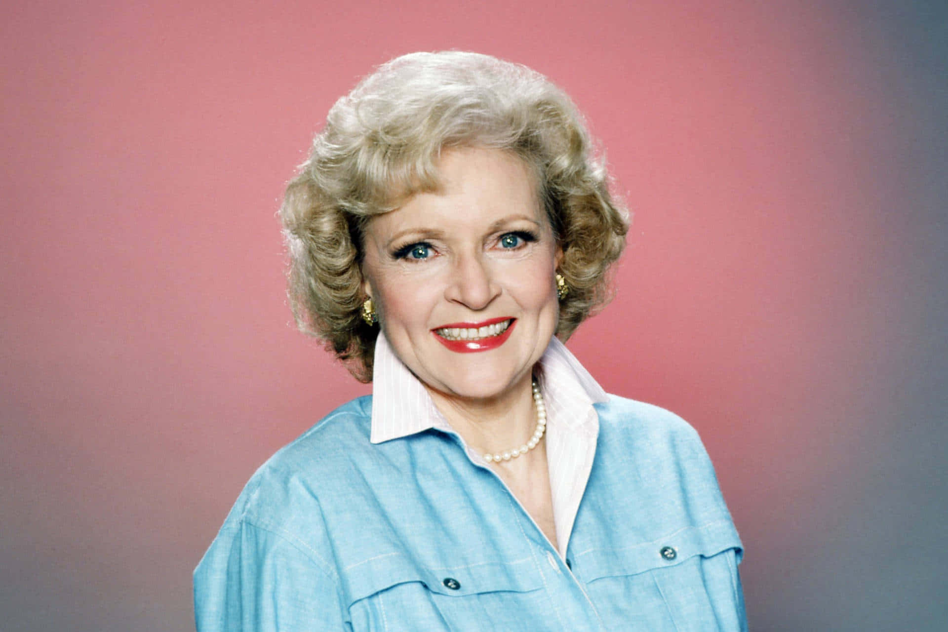 Betty White 桌布 壁紙 — 一位開朗的金髮捲曲、塗著紅色唇膏的女性，在柔和的粉紅與灰色漸變背景前溫暖微笑。
