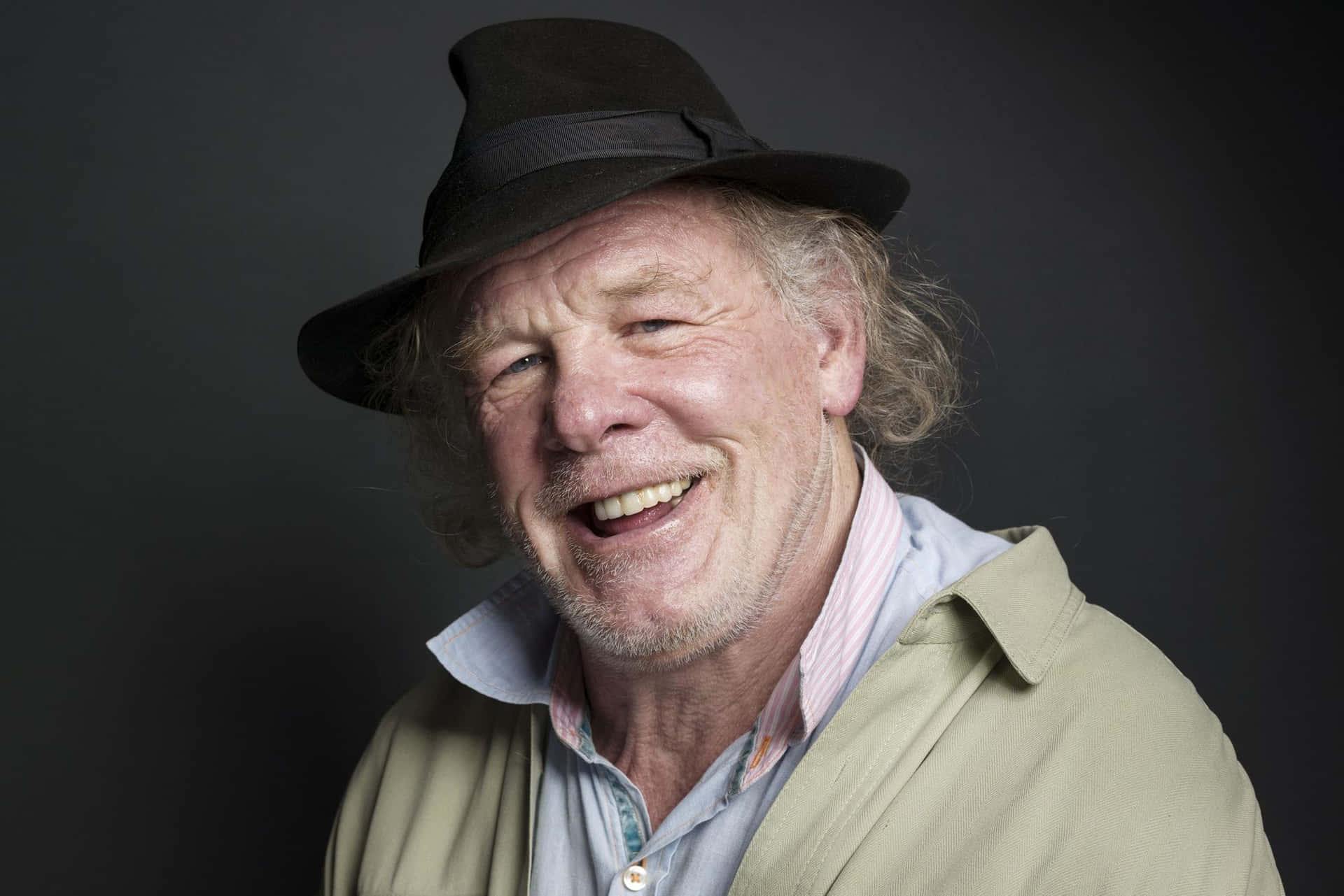 Nick Nolte 桌布 壁紙 — 一位開朗的長者，捲髮戴著軟呢帽，在深色攝影棚背景下溫暖地笑著。