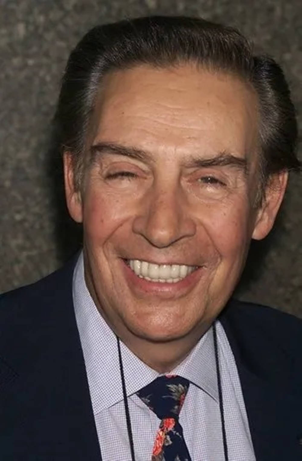 actor 桌布 壁紙 — 一位開朗的黑髮年長男士，身穿深色西裝和花卉領帶，在紋理灰色背景前燦爛微笑。
