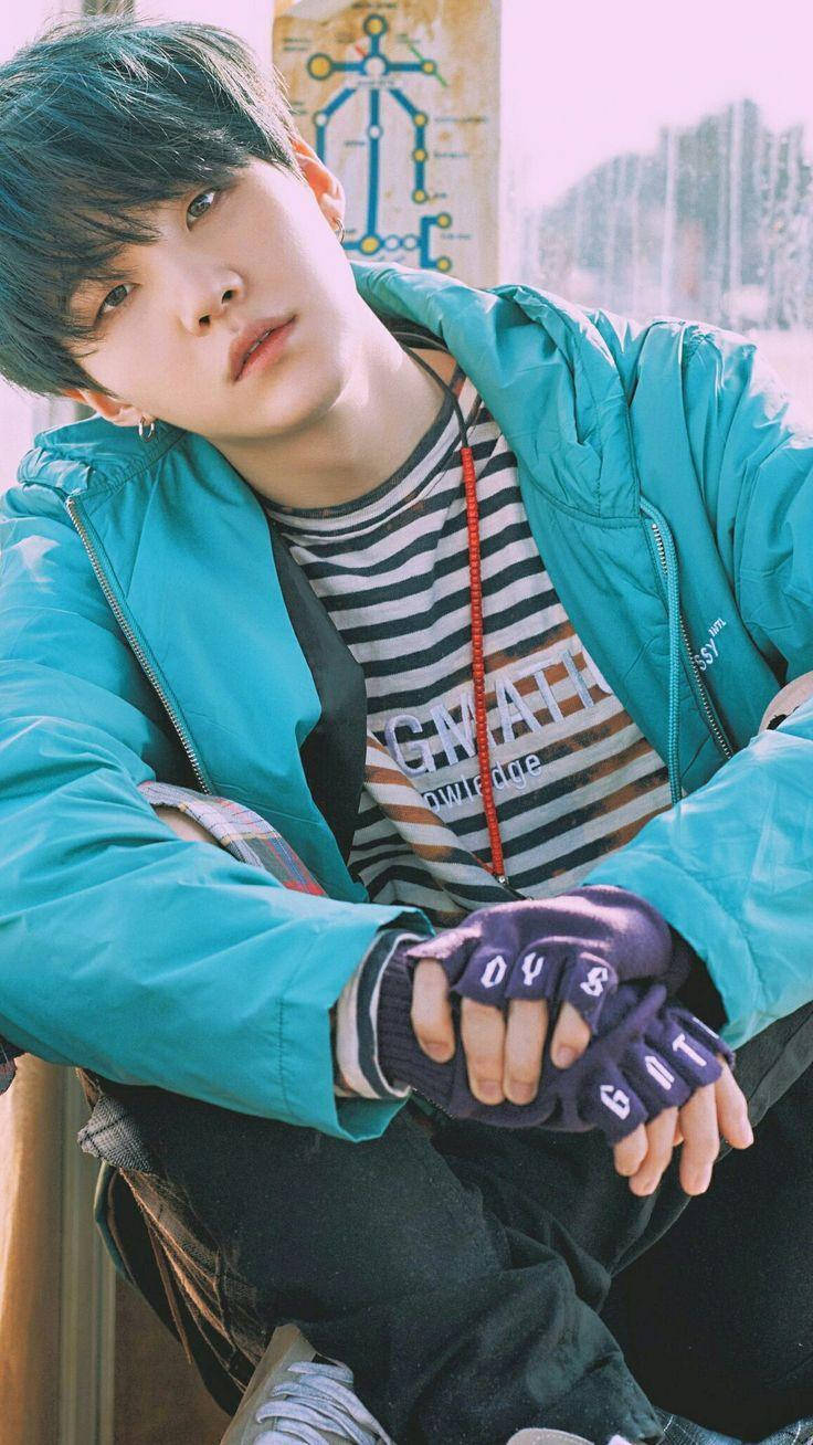 Suga Bts 桌布 壁紙 — 一位青綠色頭髮的年輕男子靠著牆，身穿亮藍色外套和無指手套。