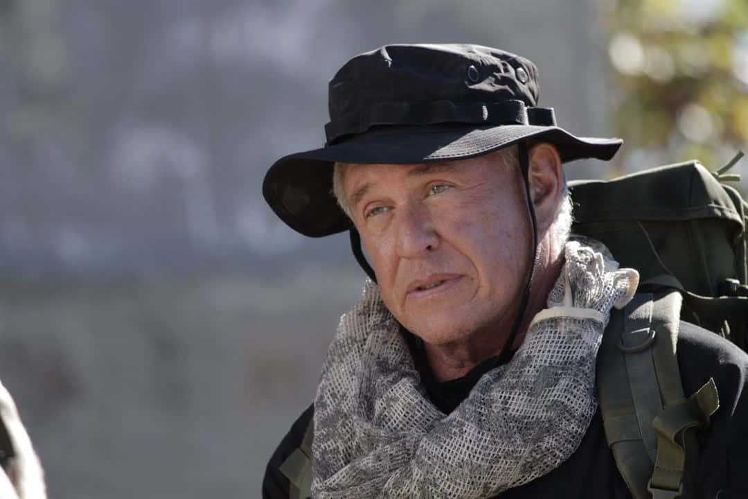 Tom Berenger 桌布 壁紙 — 一位面容滄桑的老年男子，戴著黑色奔尼帽，背著綠色背包，眺望遠方。
