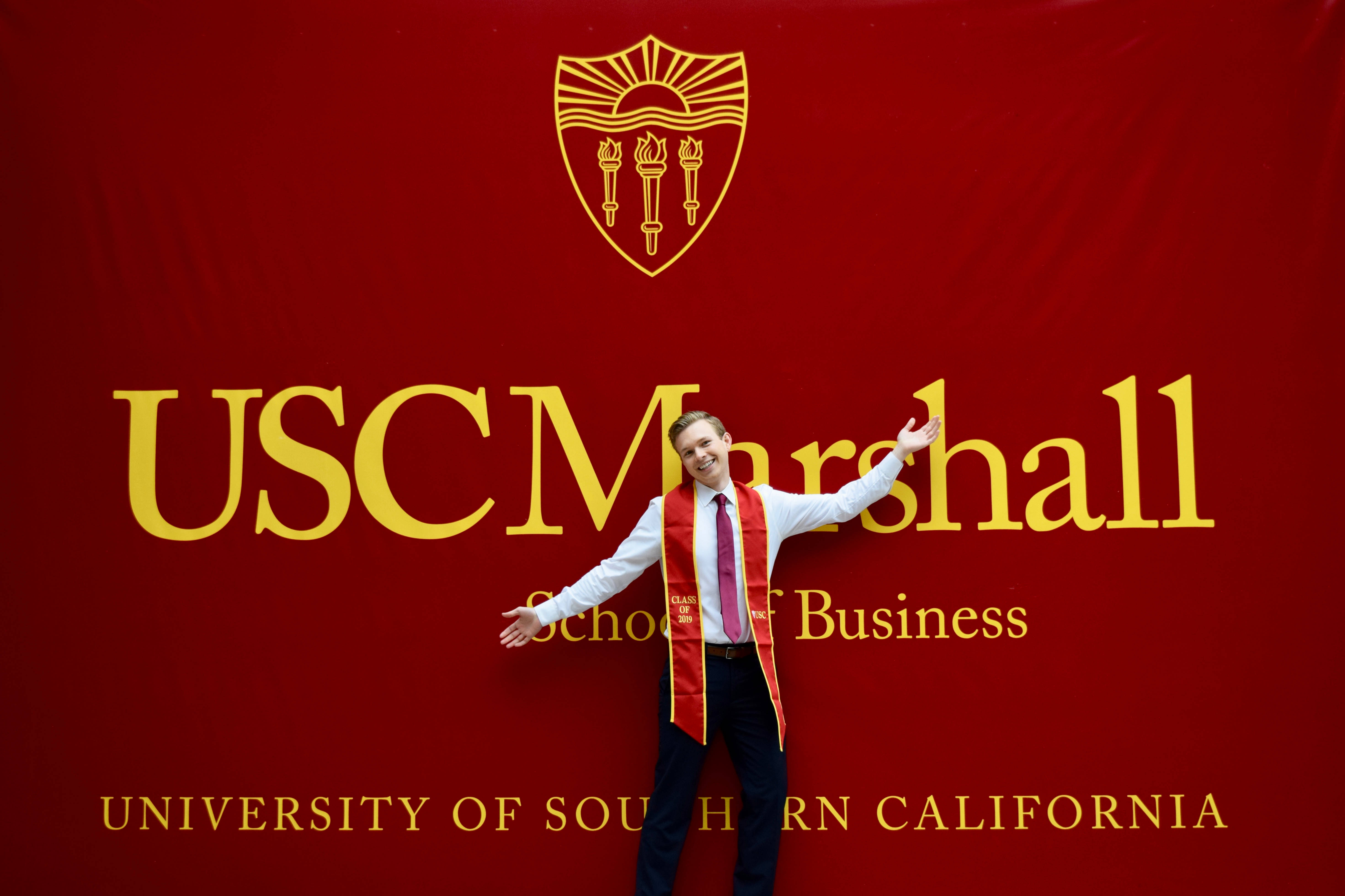 Usc 桌布 壁紙 — 一位面帶微笑、佩戴紅色肩帶的畢業生，在鮮紅色的南加州大學馬歇爾商學院背景前張開雙臂站立。
