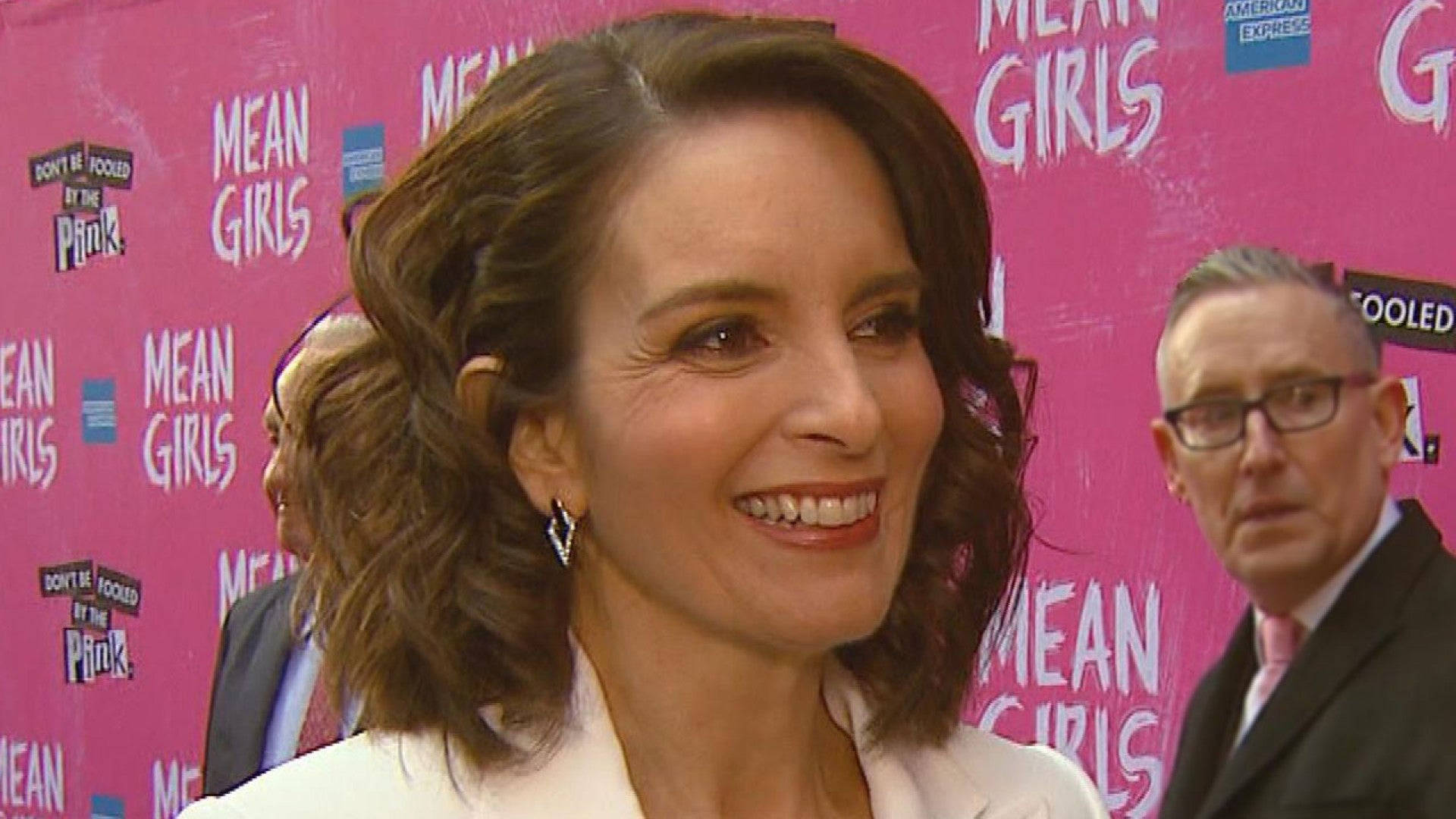 mean girls 桌布 壁紙 — 一位面帶微笑、擁有波浪棕色長髮的女性，在紅毯活動中站在明亮的粉紅色「Mean Girls」品牌背景前。
