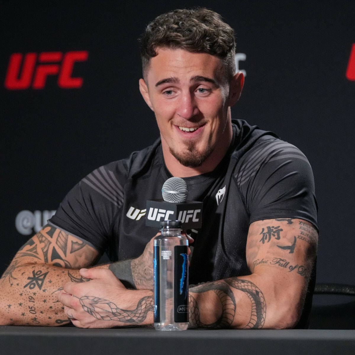 UFC 桌布 壁紙 — 一位面帶微笑、有刺青的UFC拳手坐在記者會的桌子前，桌上放著麥克風和水瓶。