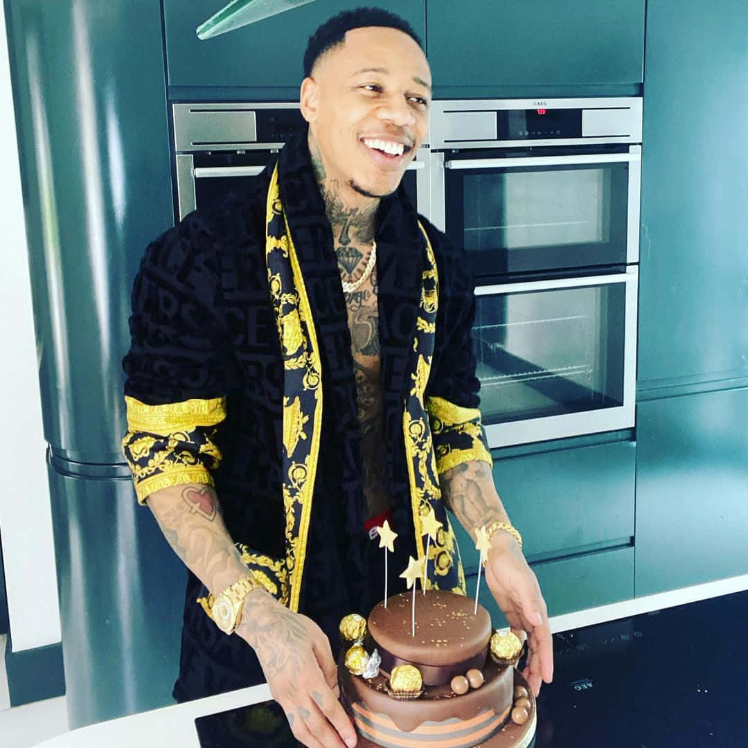 Celebrity 桌布 壁紙 — 一位面帶微笑、有刺青的男士穿著黑金色長袍，在現代廚房裡展示巧克力蛋糕。