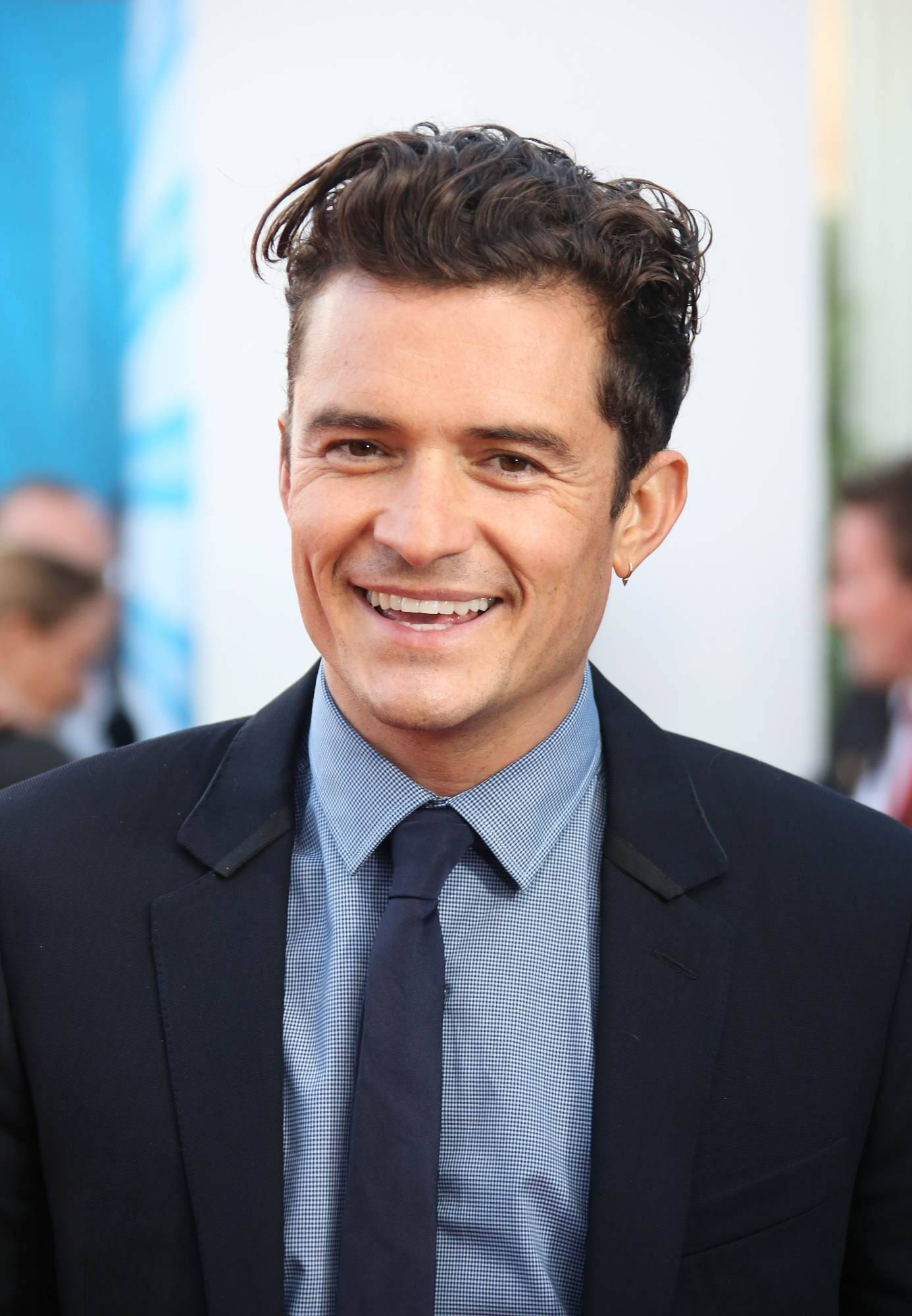 orlando bloom 桌布 壁紙 — 一位面帶微笑、梳著深色時尚髮型的男士，穿著俐落的黑色西裝和藍色格紋襯衫，背景是模糊的活動場景。