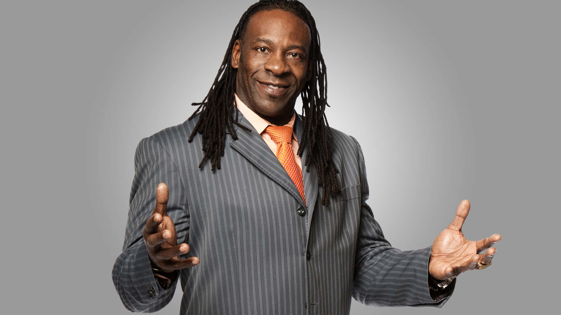 WWE 桌布 壁紙 — 一位面帶微笑、梳著辮子的男士，身穿灰色細條紋西裝和橙色領帶，在純灰色背景前張開雙手做手勢。