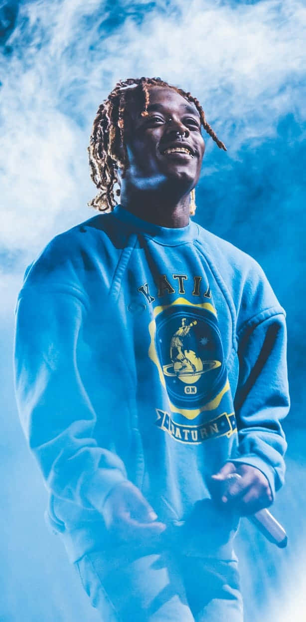  壁紙 — 一位面帶微笑、梳著辮子的男士，身穿藍色連帽上衣，在煙霧繚繞的藍色舞台燈光下拿著麥克風。