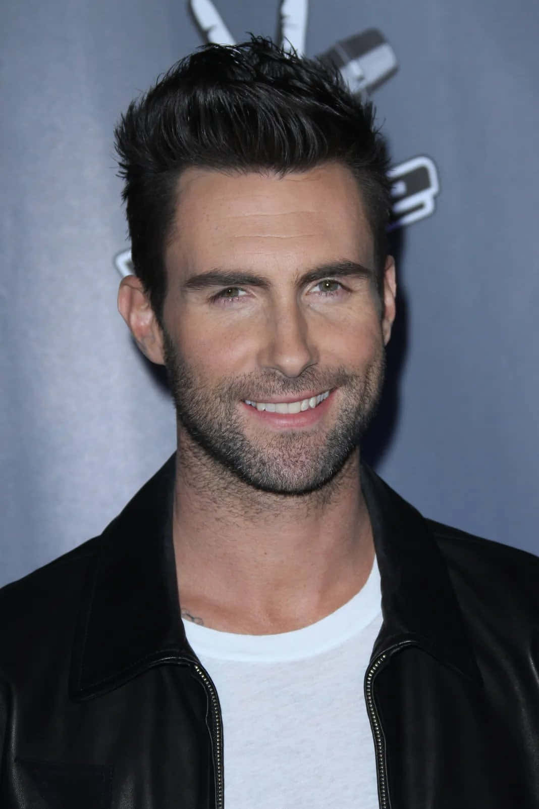 Adam Levine 桌布 壁紙 — 一位面帶微笑、深色頭髮、鬍渣的男人，在灰色背景前穿著黑色皮夾克，內搭白色T恤。