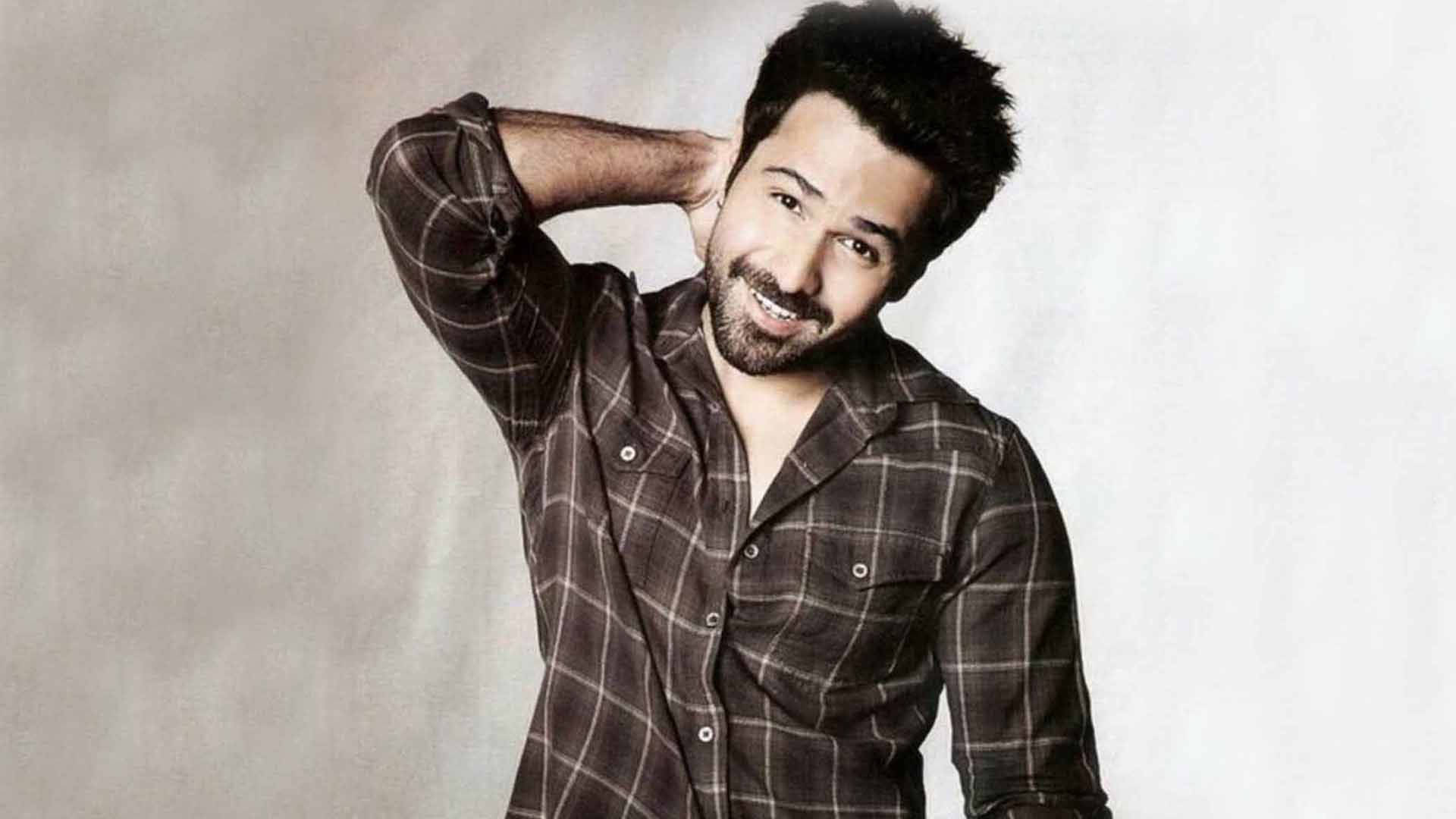 Emraan Hashmi 桌布 壁紙 — 一位面帶微笑、深色頭髮和鬍鬚的男子，穿著格子襯衫，在純色攝影棚背景前擺姿勢。