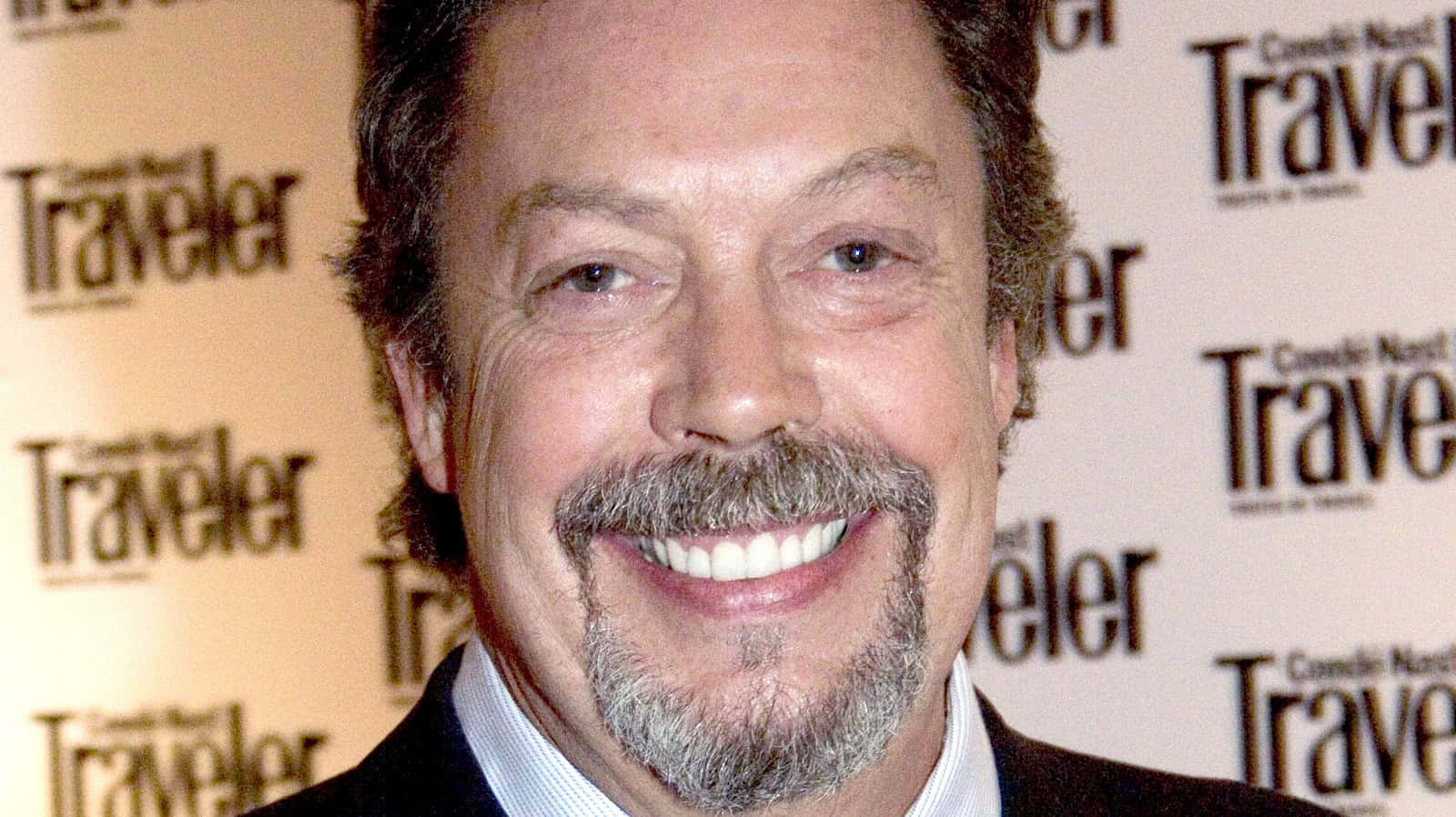 Tim Curry 桌布 壁紙 — 一位面帶微笑、留著灰白鬍鬚和鬍子的男士，在重複的Traveler雜誌背景前擺姿勢。