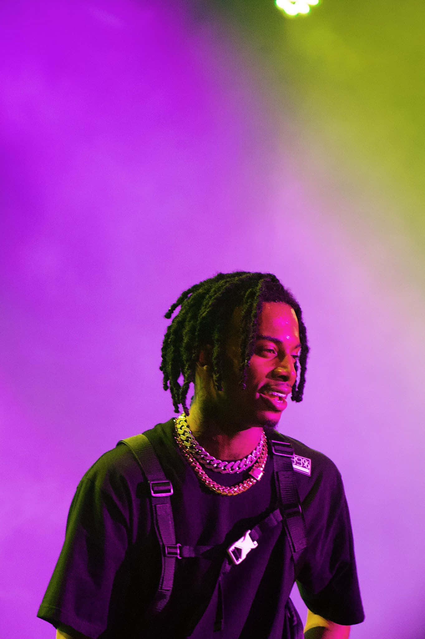Playboi Carti HD 桌布 壁紙 — 一位面帶微笑、留著辮子的男士，在紫色和綠色的舞台燈光下佩戴著多層鏈條。