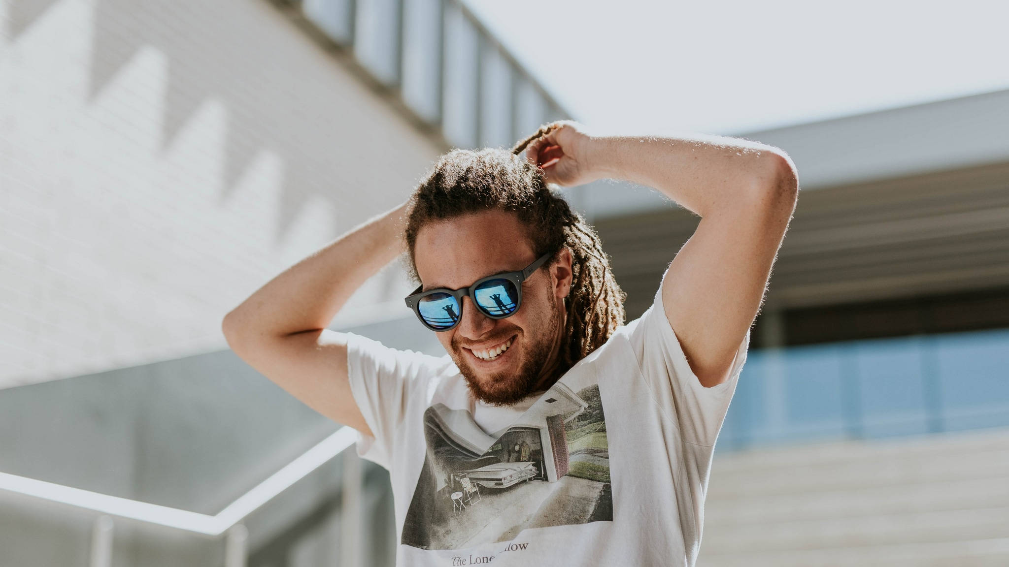 smiling 桌布 壁紙 — 一位面帶微笑、留著辮子頭並戴著太陽眼鏡的男子，在明亮的陽光下，於現代建築外牆前用手撥弄頭髮。