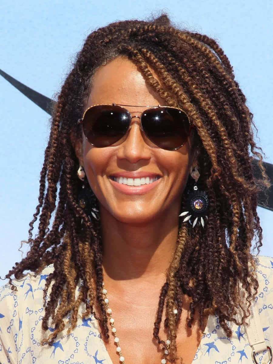 Nicole Ari Parker 桌布 壁紙 — 一位面帶微笑、留著長辮子、戴著太陽眼鏡的女性，站在晴朗的藍天下，背景是星形圖案。