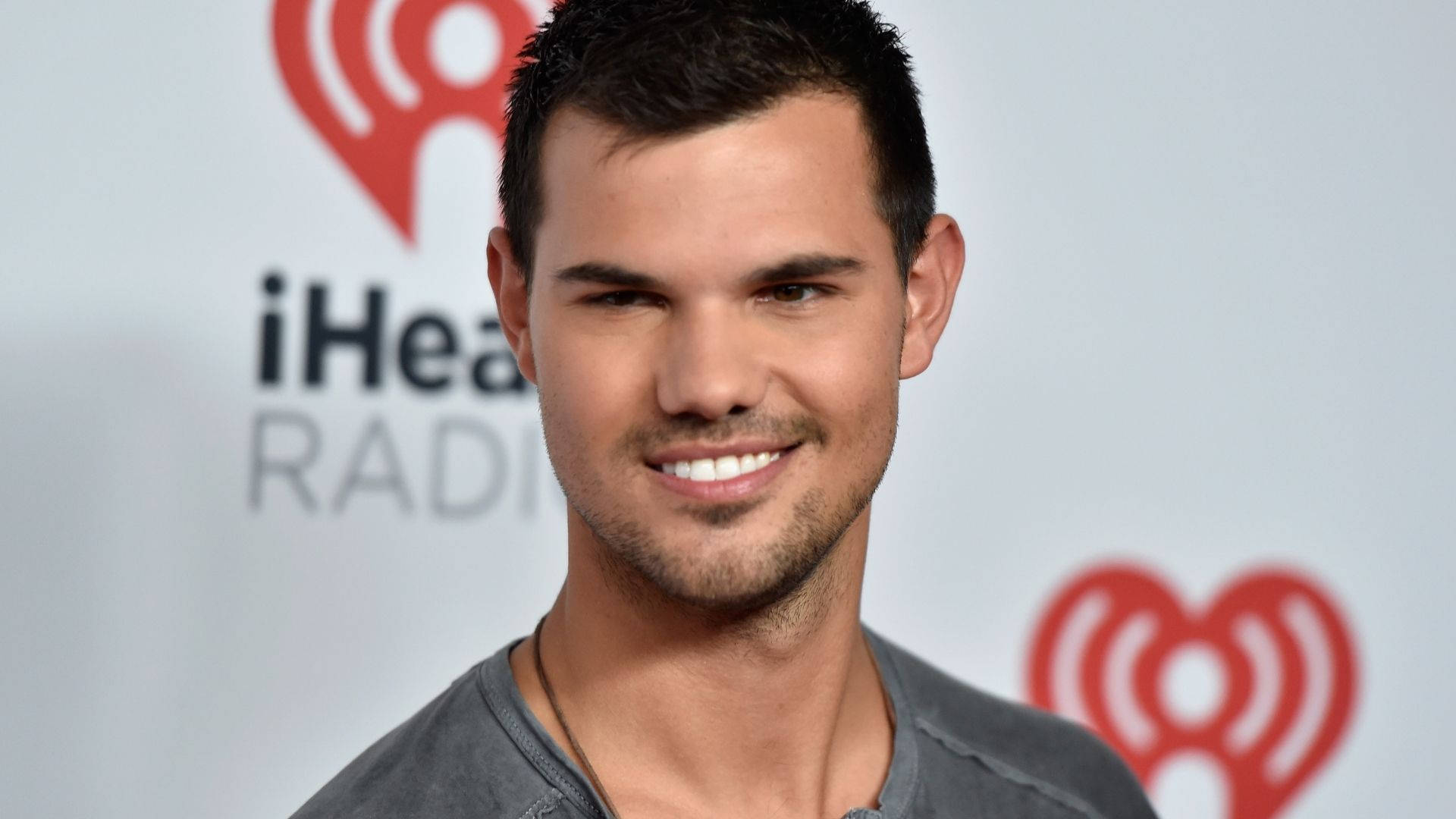 Taylor Lautner 桌布 壁紙 — 一位面帶微笑、短黑髮、有鬍渣的男子，在白色背景上帶有紅色iHeartRadio標誌的情況下穿著灰色襯衫。