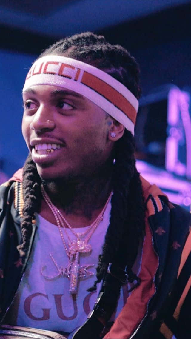  壁紙 — 一位面帶微笑、編著辮子的男士，在紫色舞台燈光下戴著Gucci頭帶和分層首飾。