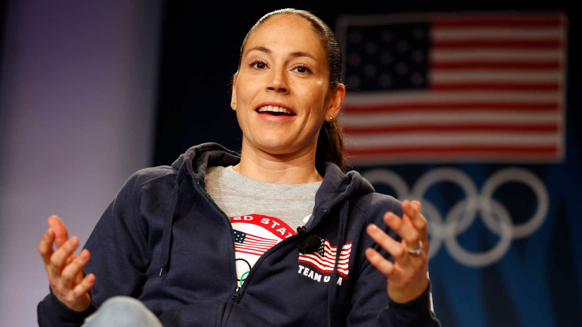 Sue 桌布 壁紙 — 一位面帶微笑、身穿Team USA連帽衫的女性，在美國國旗和奧運五環前說話並做手勢。