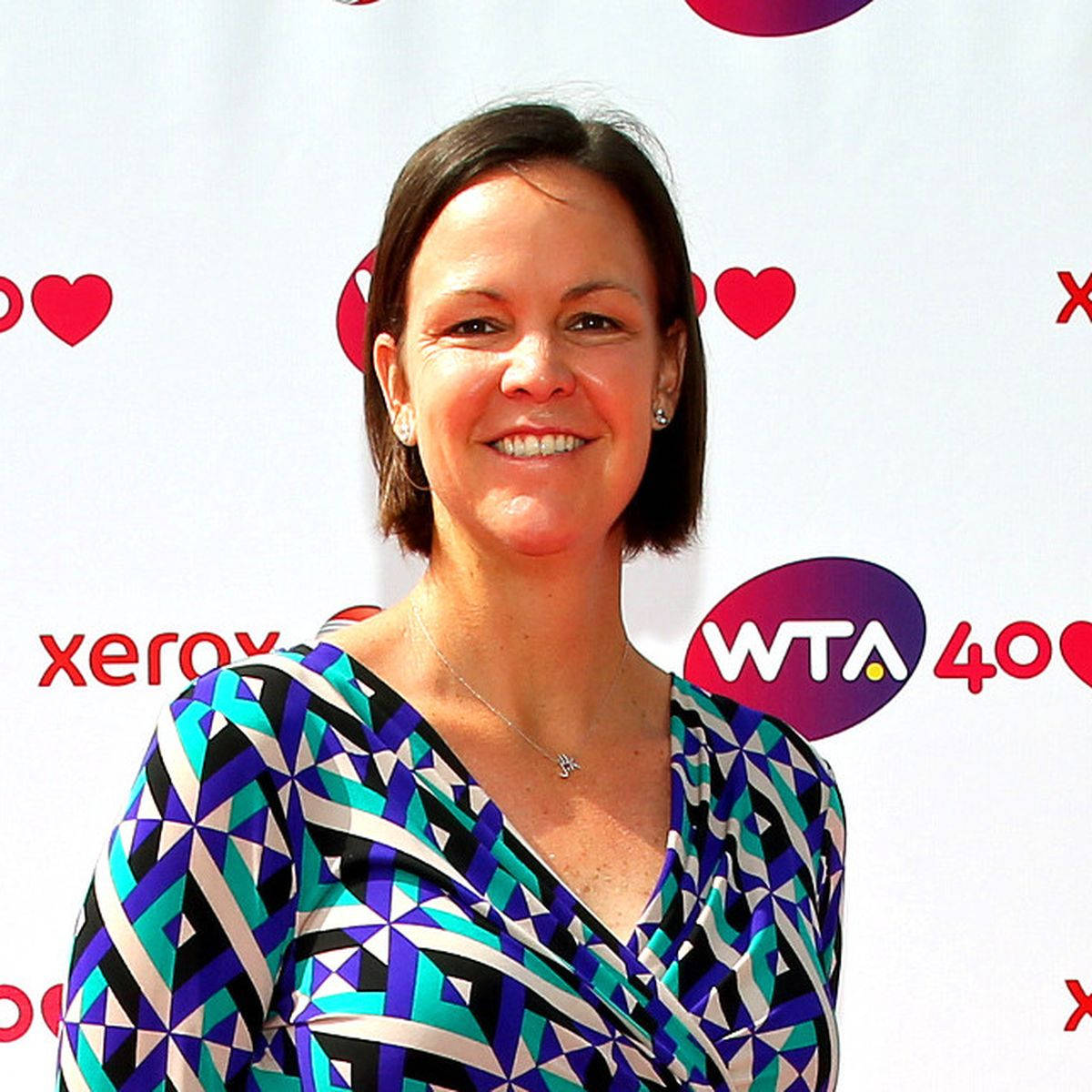 Tennis 桌布 壁紙 — 一位面帶微笑、身穿幾何圖案上衣的女性站在白色背景前，背景上有紅色心形和WTA品牌標誌。