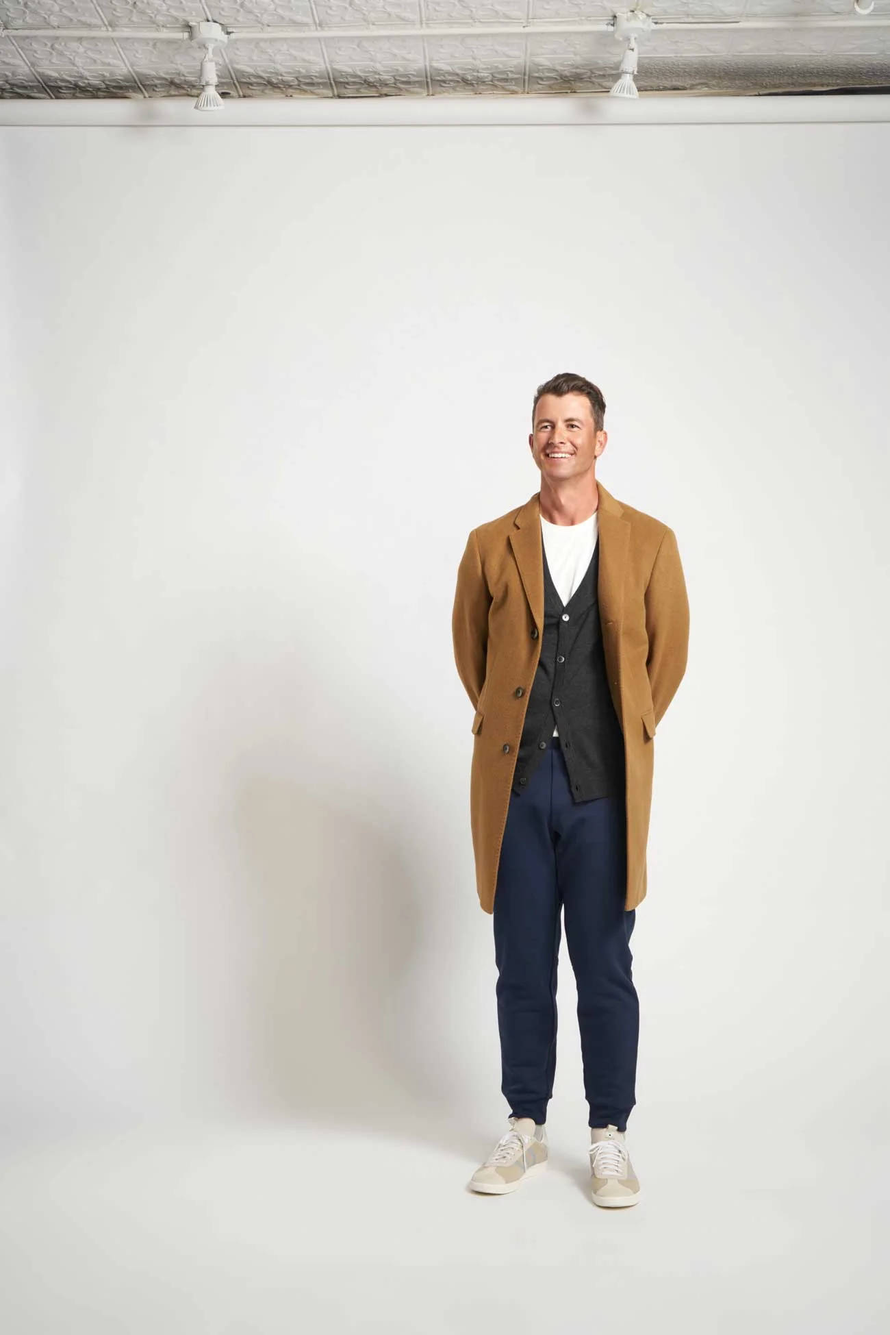 adam scott 桌布 壁紙 — 一位面帶微笑、身穿棕褐色外套和海軍藍褲子的男士，自信地站在純白色攝影棚背景前。