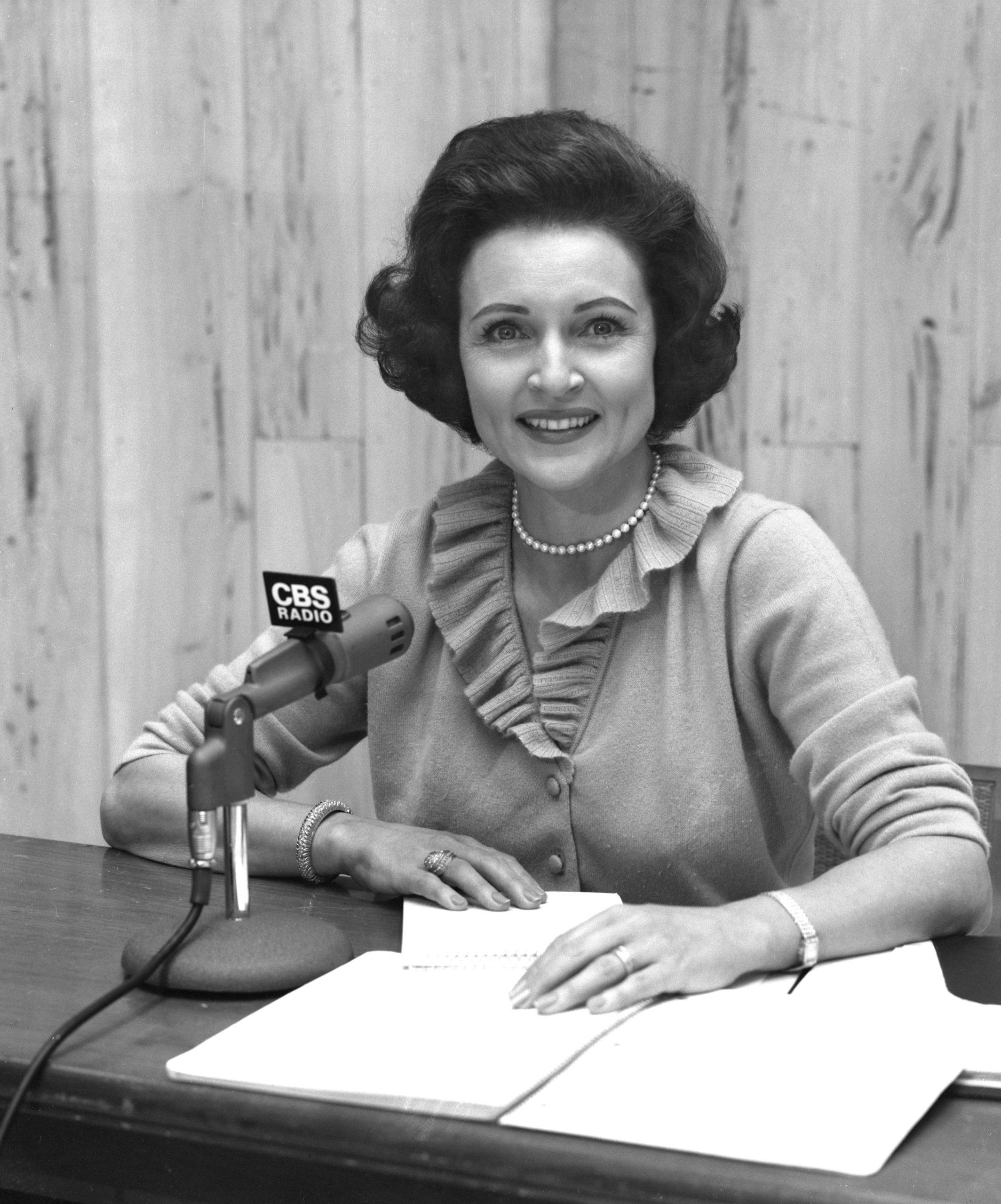 Betty White 桌布 壁紙 — 一位面帶微笑、身穿荷葉邊開襟衫的女性坐在辦公桌前，桌上放著CBS Radio麥克風和文件。