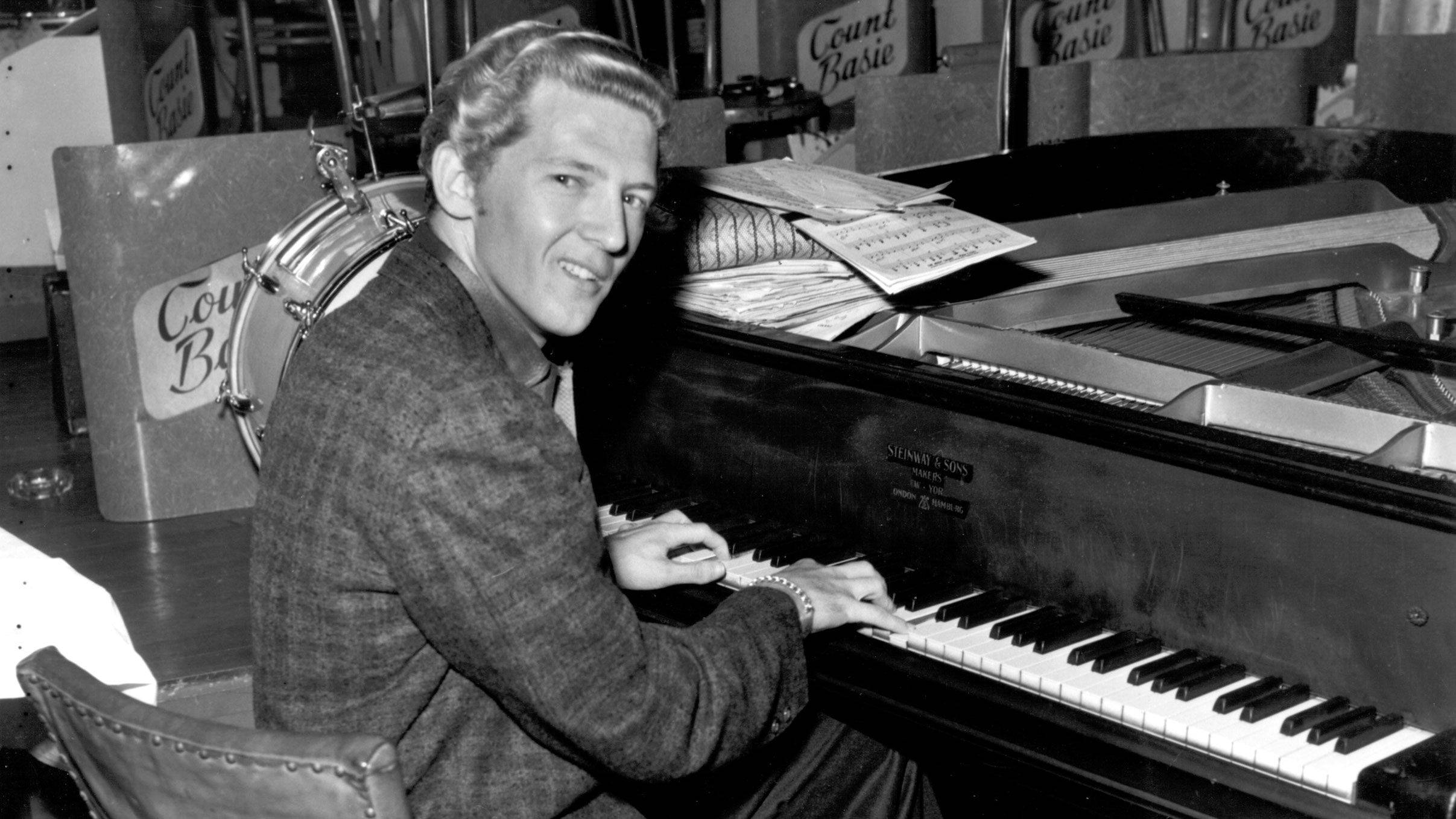 Jerry Lee Lewis 桌布 壁紙 — 一位面帶微笑、身穿質感外套的男子，在爵士俱樂部中，周圍有「Count Basie」標誌和樂譜，正在彈奏三角鋼琴。