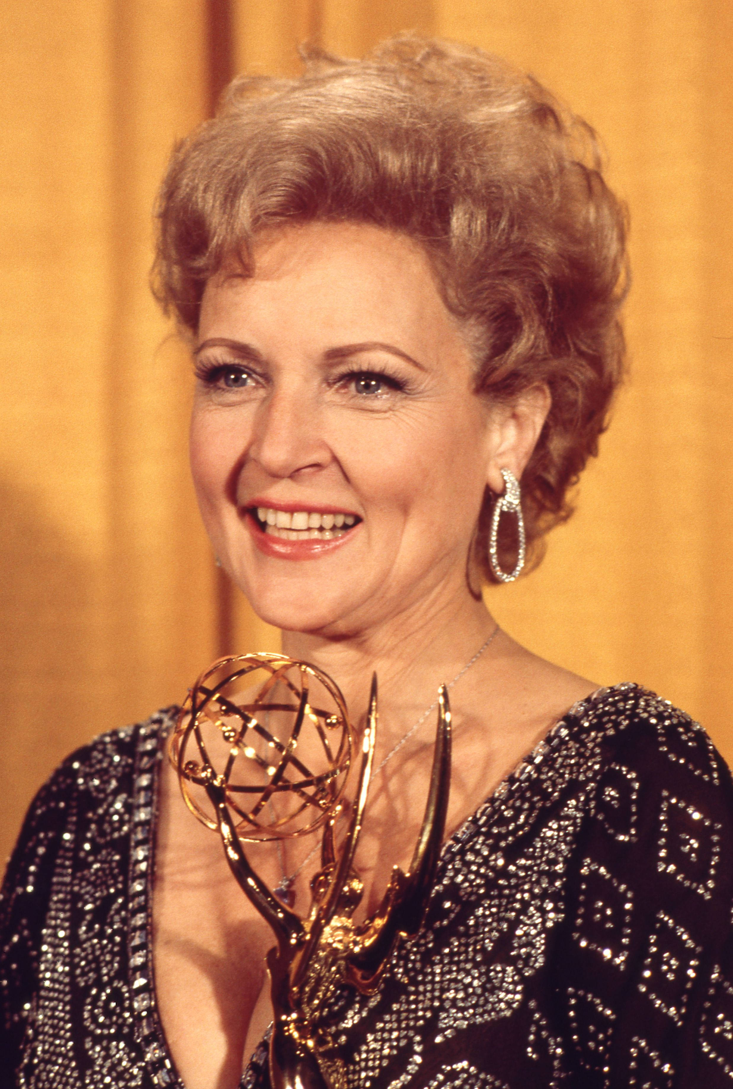 Betty White 桌布 壁紙 — 一位面帶微笑、頭髮造型時尚的女性，在溫暖的黃色窗簾背景下手持金色艾美獎獎座。