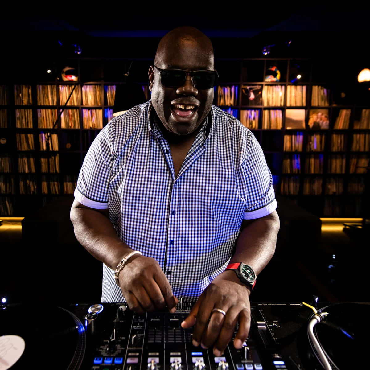 DJ 桌布 壁紙 — 一位面帶微笑的DJ穿著格紋襯衫，在昏暗的俱樂部燈光下，於帶有發光控制器的唱盤上混音。