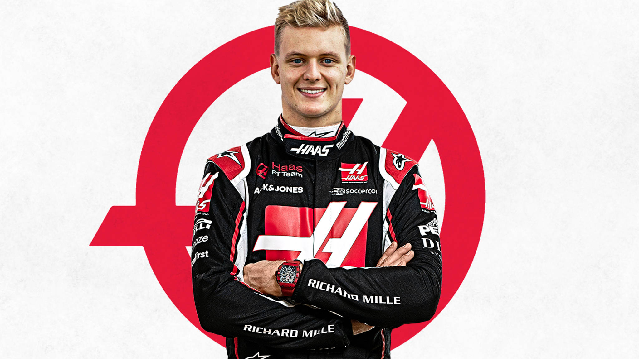 mick schumacher 桌布 壁紙 — 一位面帶微笑的F1車手身穿黑紅色的Haas賽車服，雙臂交叉，自信地站在白色背景上一個醒目的紅色圓形標誌前。