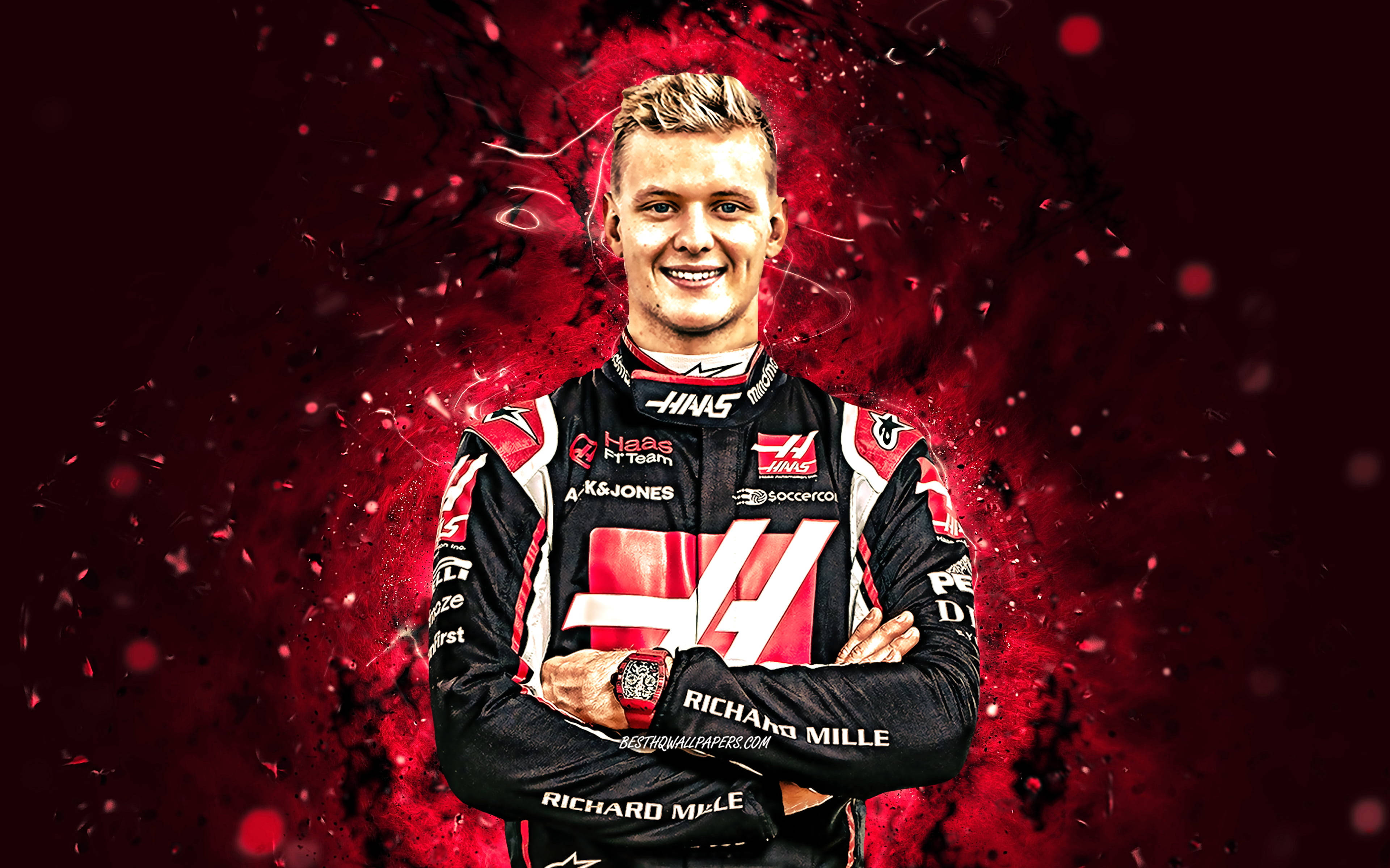 mick schumacher 桌布 壁紙 — 一位面帶微笑的F1車手身穿黑紅色Haas賽車服，雙臂交叉，自信地站在動態紅色粒子背景前。