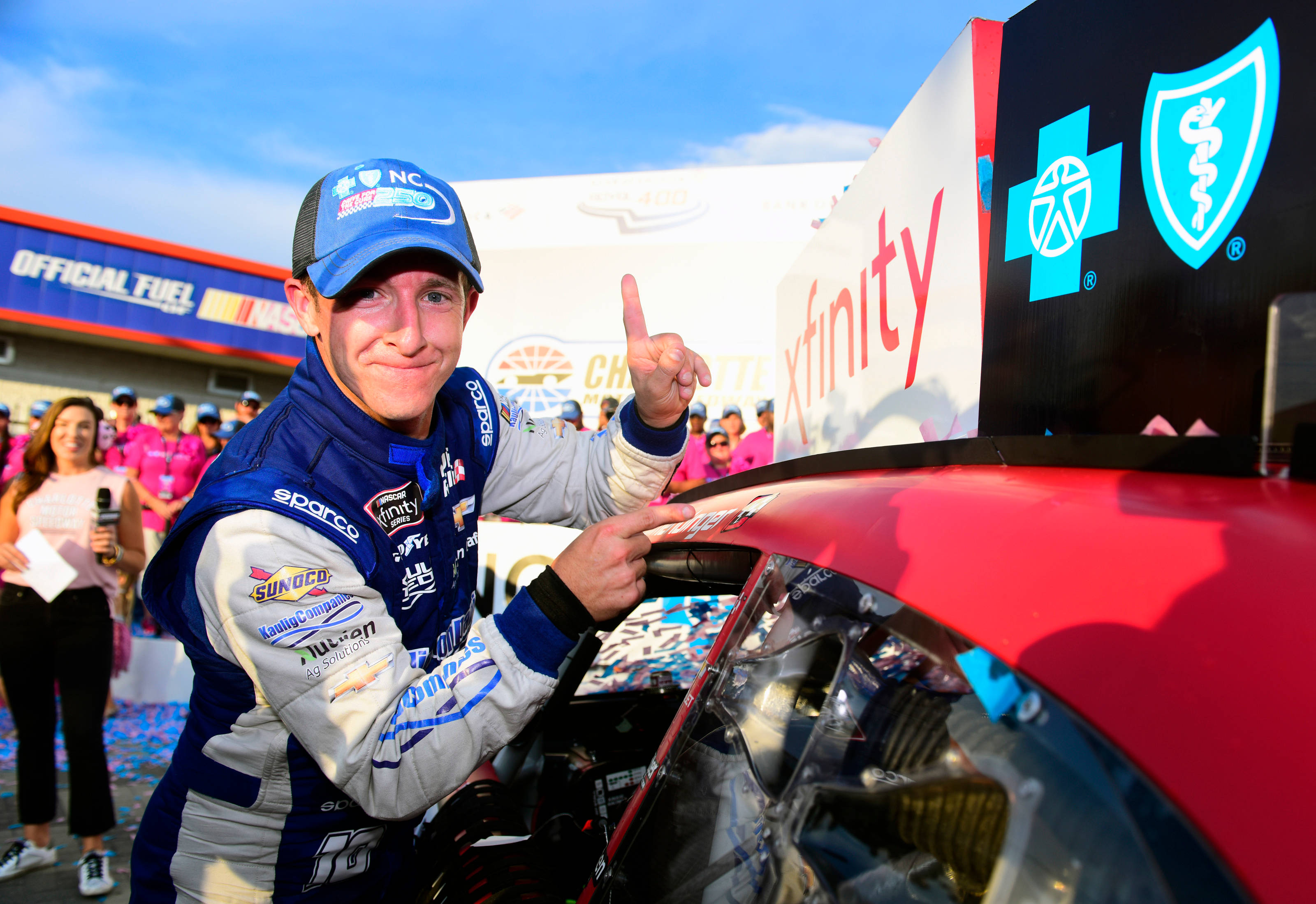 Motorsport 桌布 壁紙 — 一位面帶微笑的NASCAR車手，身穿藍白賽車服，在陽光明媚的維修站慶祝活動中，得意地指向他的紅色賽車。