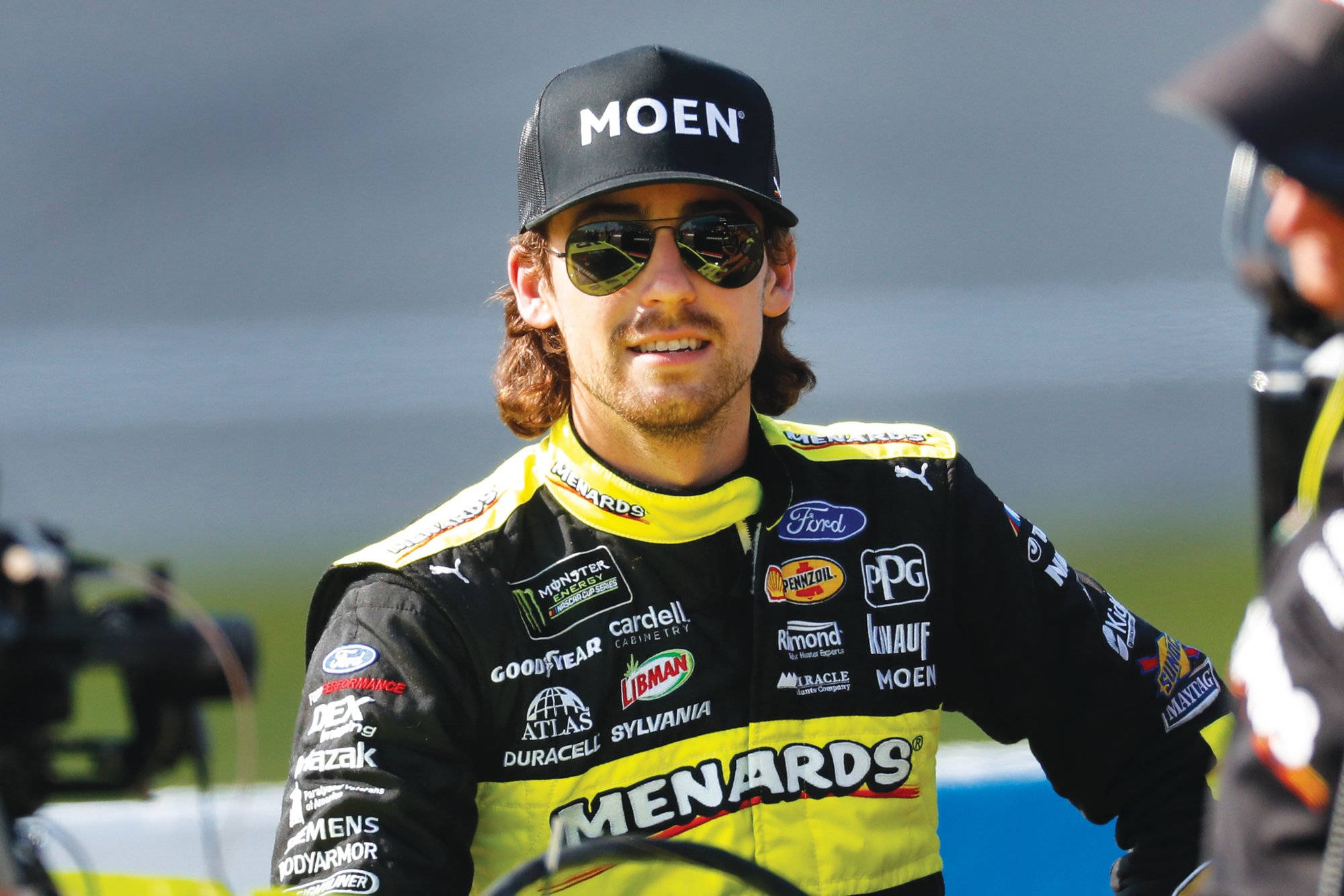 Ryan Blaney 桌布 壁紙 — 一位面帶微笑的NASCAR車手，身穿黑黃相間的Menards賽車服和帽子，站在戶外，附近有攝影機。
