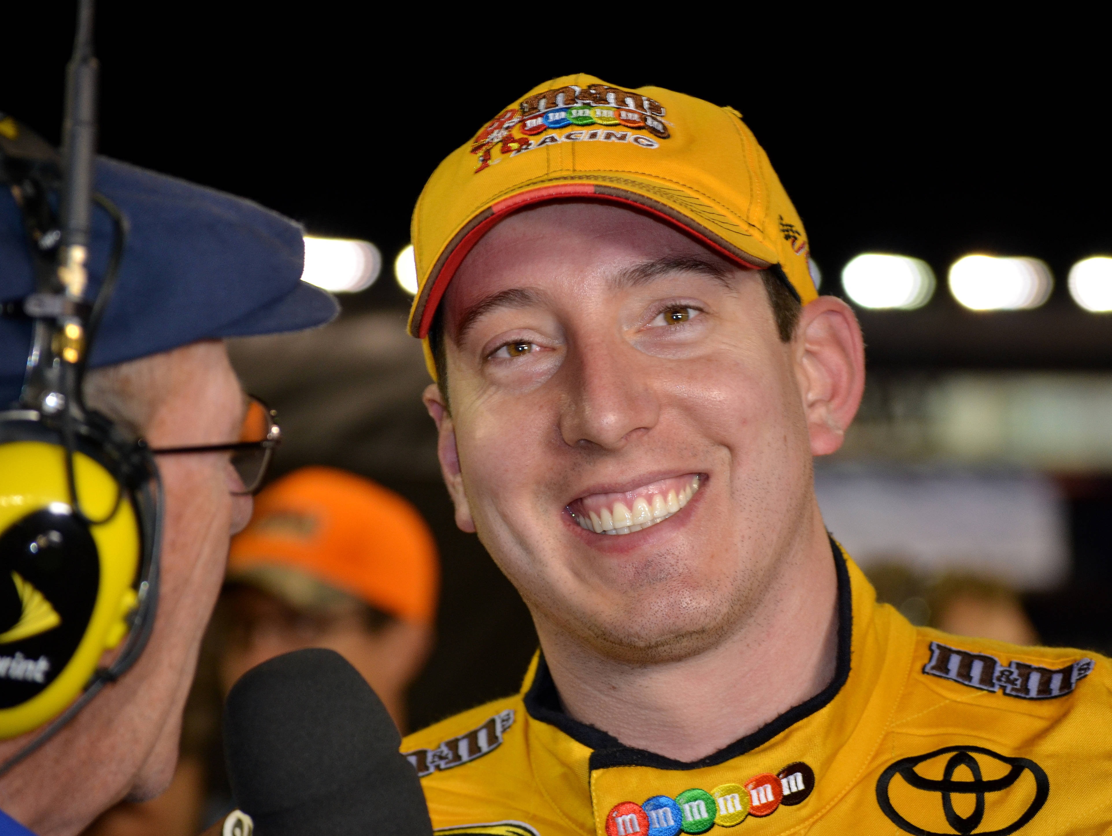 Smile 桌布 壁紙 — 一位面帶微笑的NASCAR車手穿著黃色M&M's Toyota制服，在夜晚明亮的體育場燈光下與記者聊天。