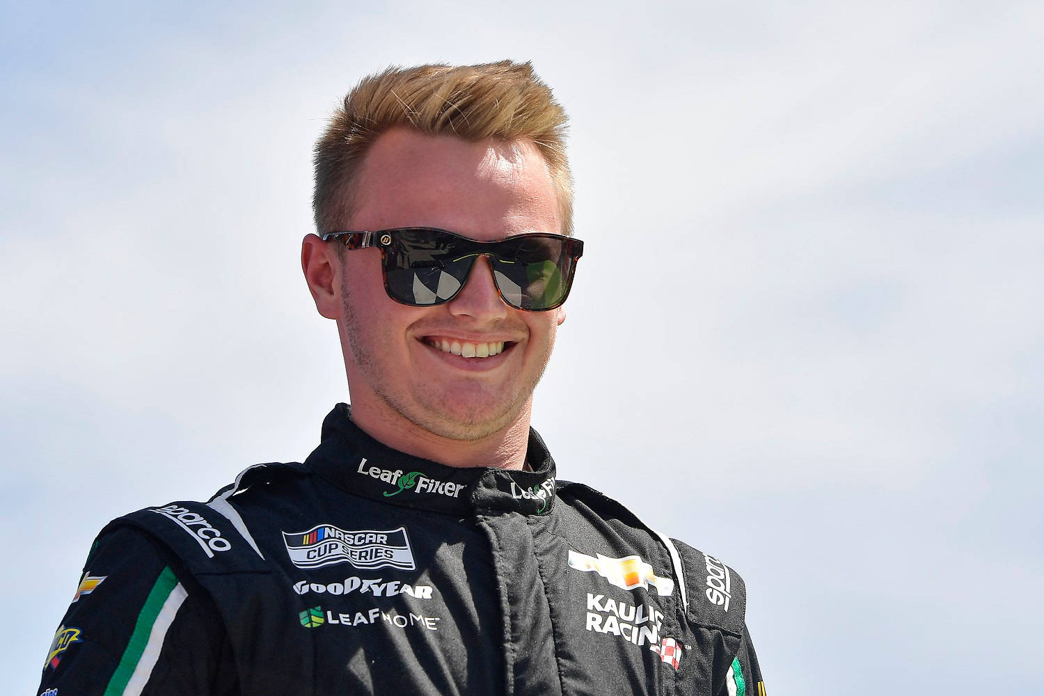 Sunglasses 桌布 壁紙 — 一位面帶微笑的NASCAR車手身穿帶有贊助商標誌的黑色賽車服，站在明亮的天空下。