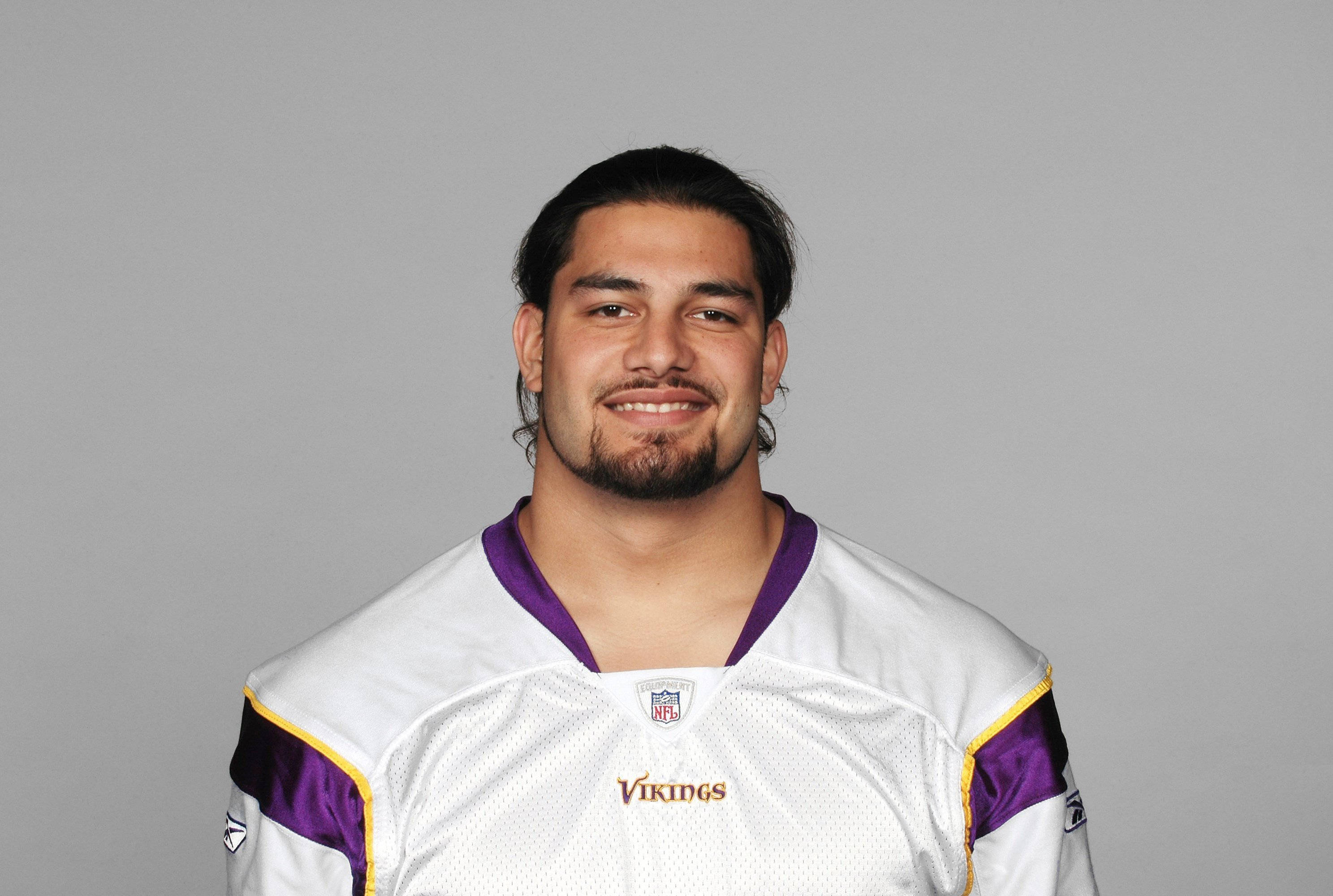 Roman 桌布 壁紙 — 一位面帶微笑的NFL美式足球員，身穿白色、紫色和金色的維京人球衣，在純灰色攝影棚背景前擺姿勢。