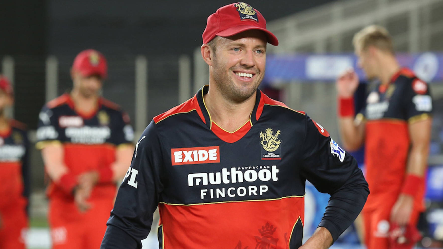 royal challengers bangalore 桌布 壁紙 — 一位面帶微笑的板球運動員，身穿帶有贊助商標誌的紅黑色制服，自信地走在體育場的場地上。