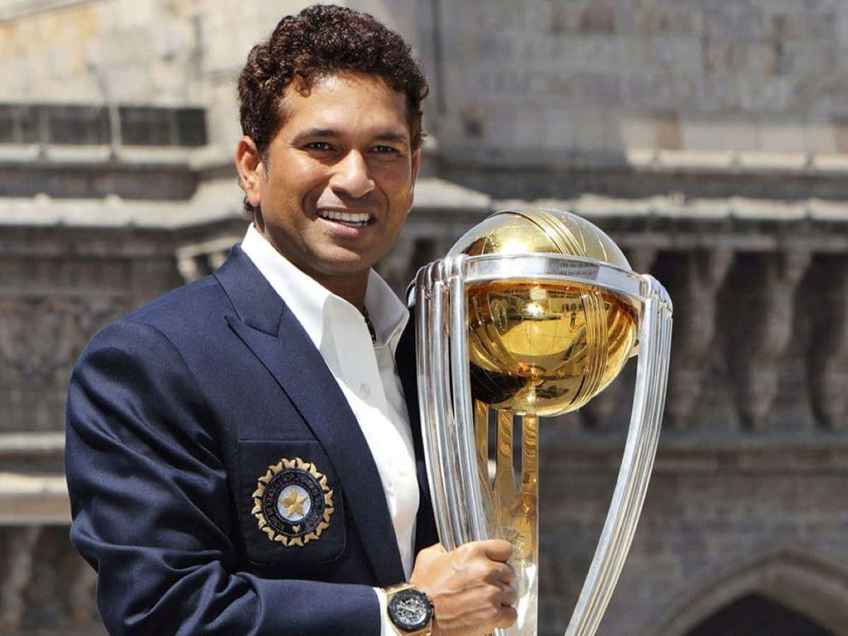Sachin Tendulkar 桌布 壁紙 — 一位面帶微笑的板球運動員身穿海軍藍西裝外套，在歷史悠久的石建築背景下，手持閃閃發光的金色板球獎盃。