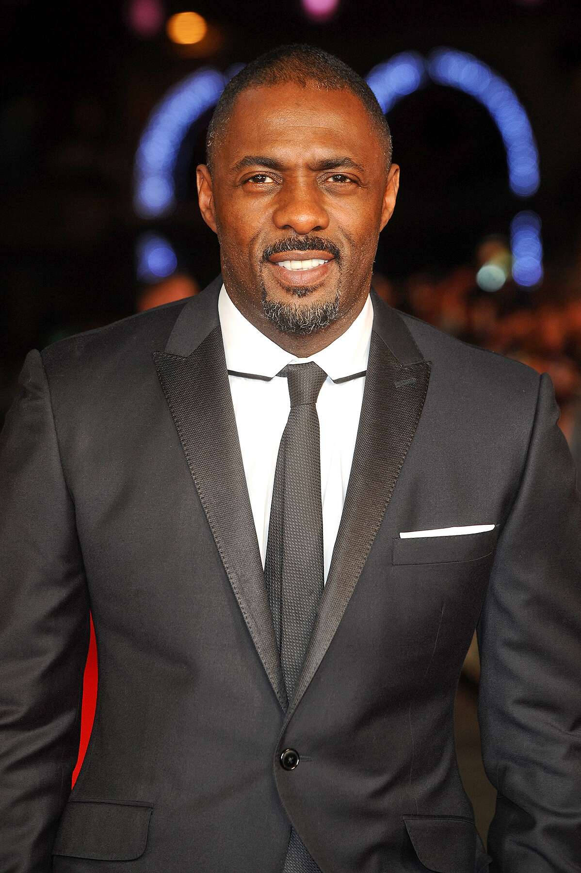 Celebrity 桌布 壁紙 — 一位面帶微笑的男士穿著剪裁俐落的黑色燕尾服，搭配緞面翻領，在模糊的夜晚背景和發光的藍色燈光下自信地站立。
