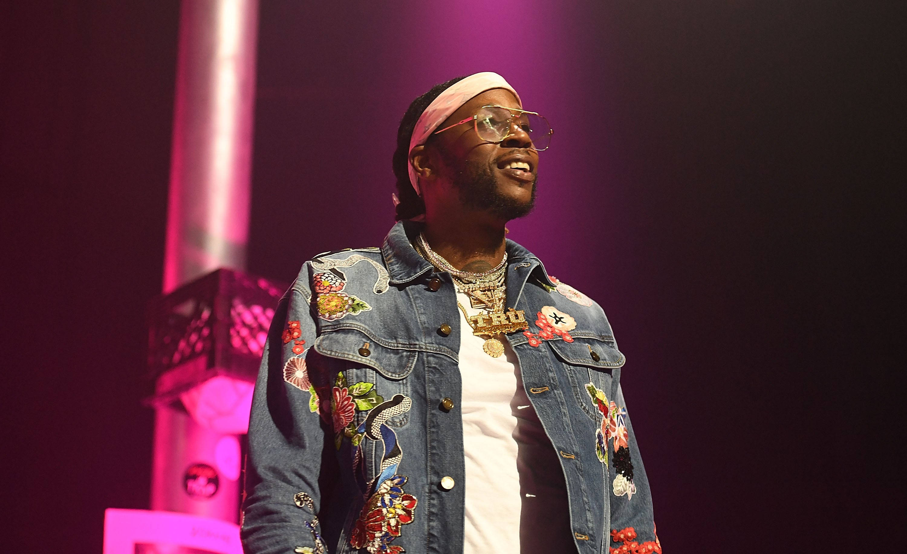 2 Chainz 桌布 壁紙 — 一位面帶微笑的男子身穿花卉圖案牛仔外套和眼鏡，在充滿活力的粉紅色舞台燈光下表演。
