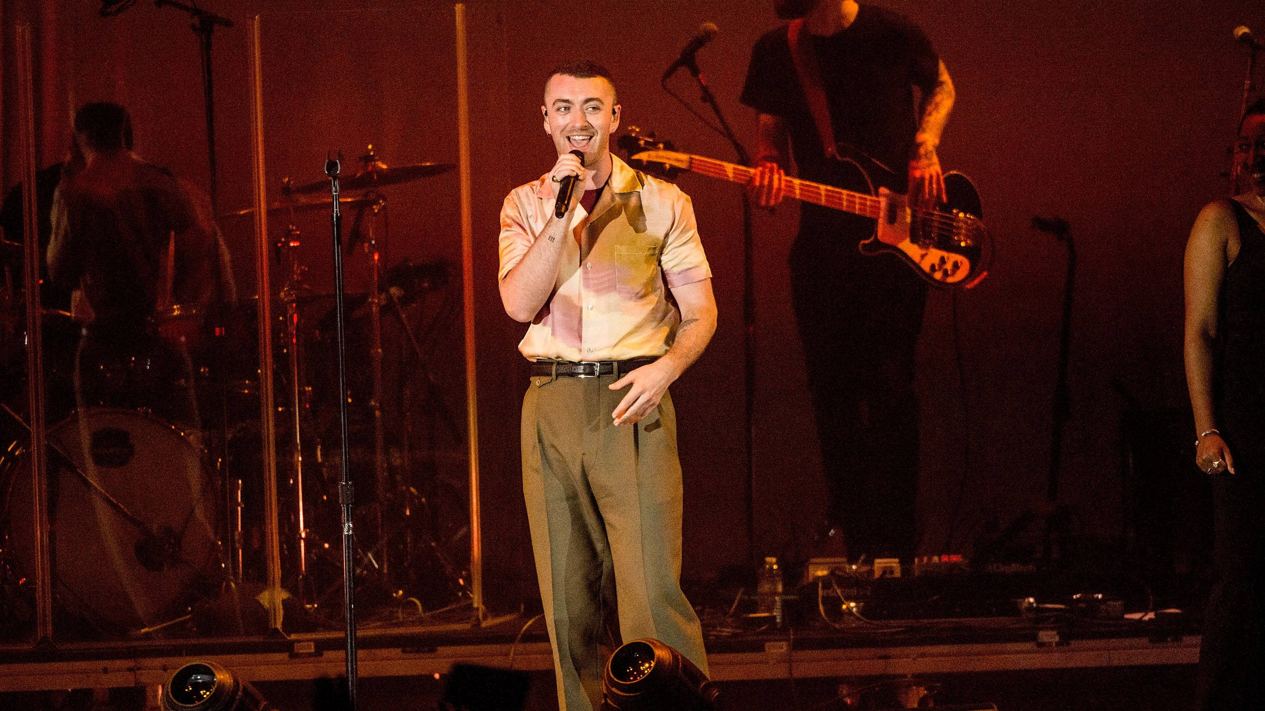 Sam Smith 桌布 壁紙 — 一位面帶微笑的男歌手身穿圖案襯衫，在溫暖燈光的舞台上表演，身後有樂隊成員和樂器。