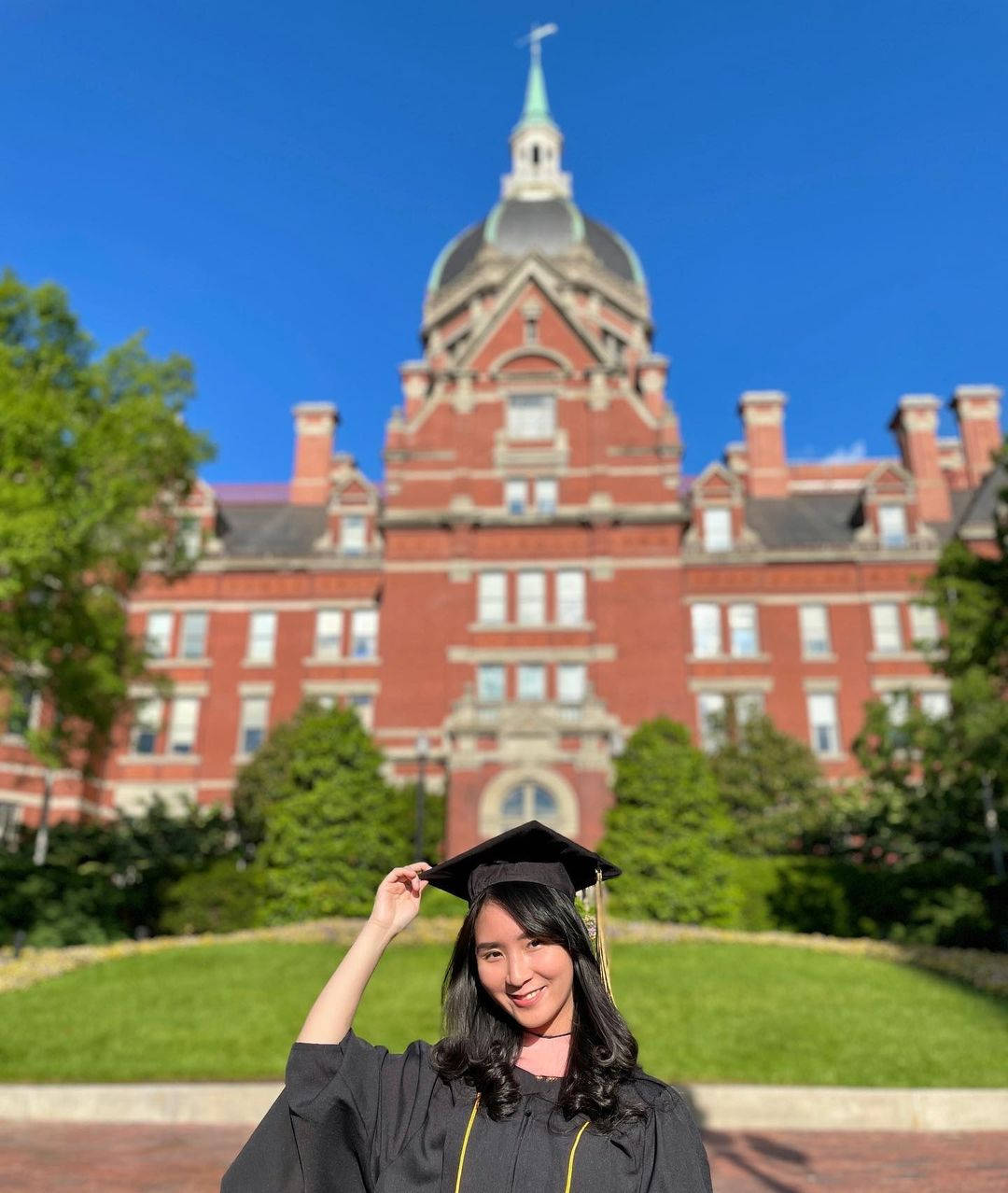 college 桌布 壁紙 — 一位面帶微笑的畢業生身穿黑色畢業袍和學士帽，在晴朗藍天下，於宏偉的紅磚大學建築前頑皮地傾斜帽子。