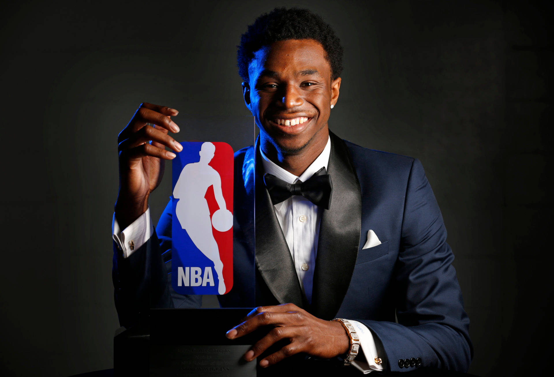 Andrew Wiggins 桌布 壁紙 — 一位面帶微笑的籃球運動員穿著俐落的藍色燕尾服，在深色攝影棚背景下自豪地高舉NBA獎盃。