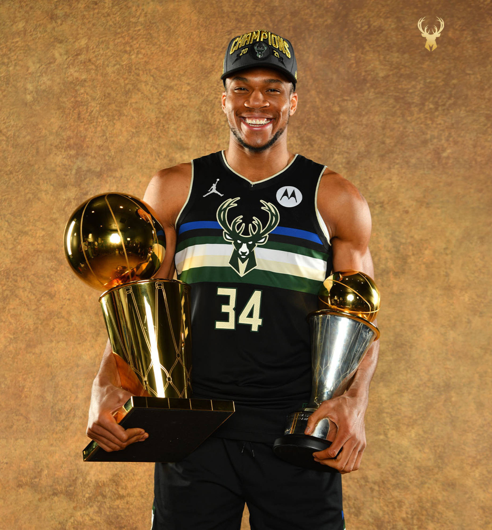 Giannis 桌布 壁紙 — 一位面帶微笑的籃球運動員身穿密爾瓦基公鹿隊球衣，在紋理金色背景前手持兩個閃閃發光的獎盃。