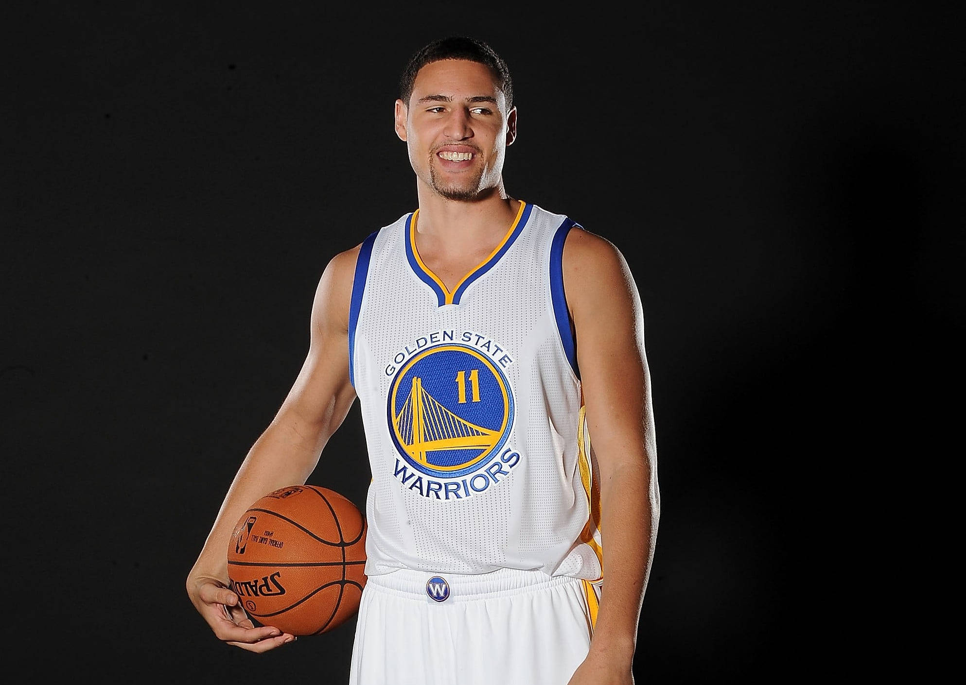 Thompson 桌布 壁紙 — 一位面帶微笑的籃球運動員身穿金州勇士隊球衣，在黑色背景前手持 Spalding 籃球。