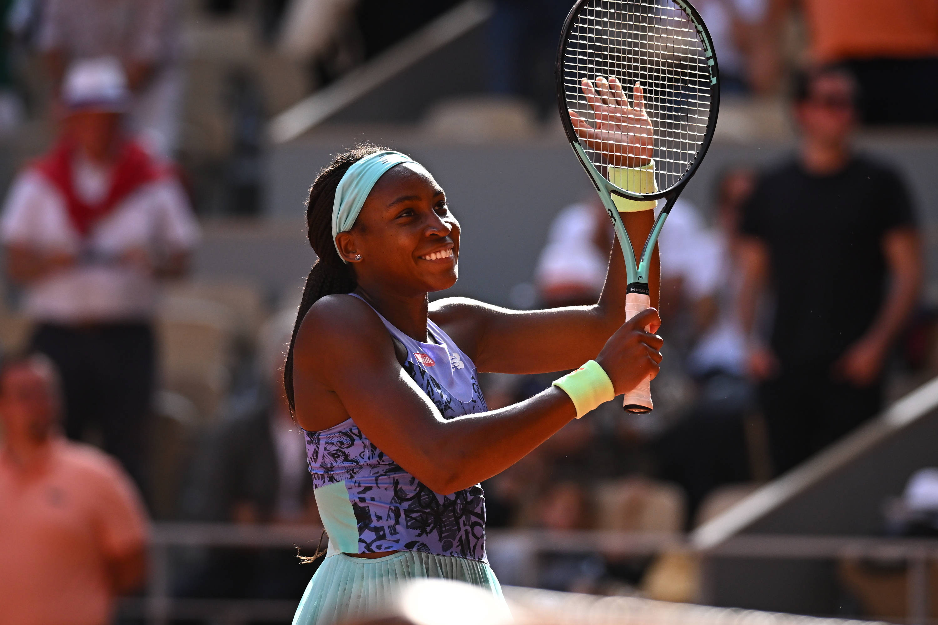 Coco Gauff 桌布 壁紙 — 一位面帶微笑的網球選手身穿圖案服裝，在陽光明媚的紅土場上高舉球拍慶祝勝利，身後是模糊的觀眾。