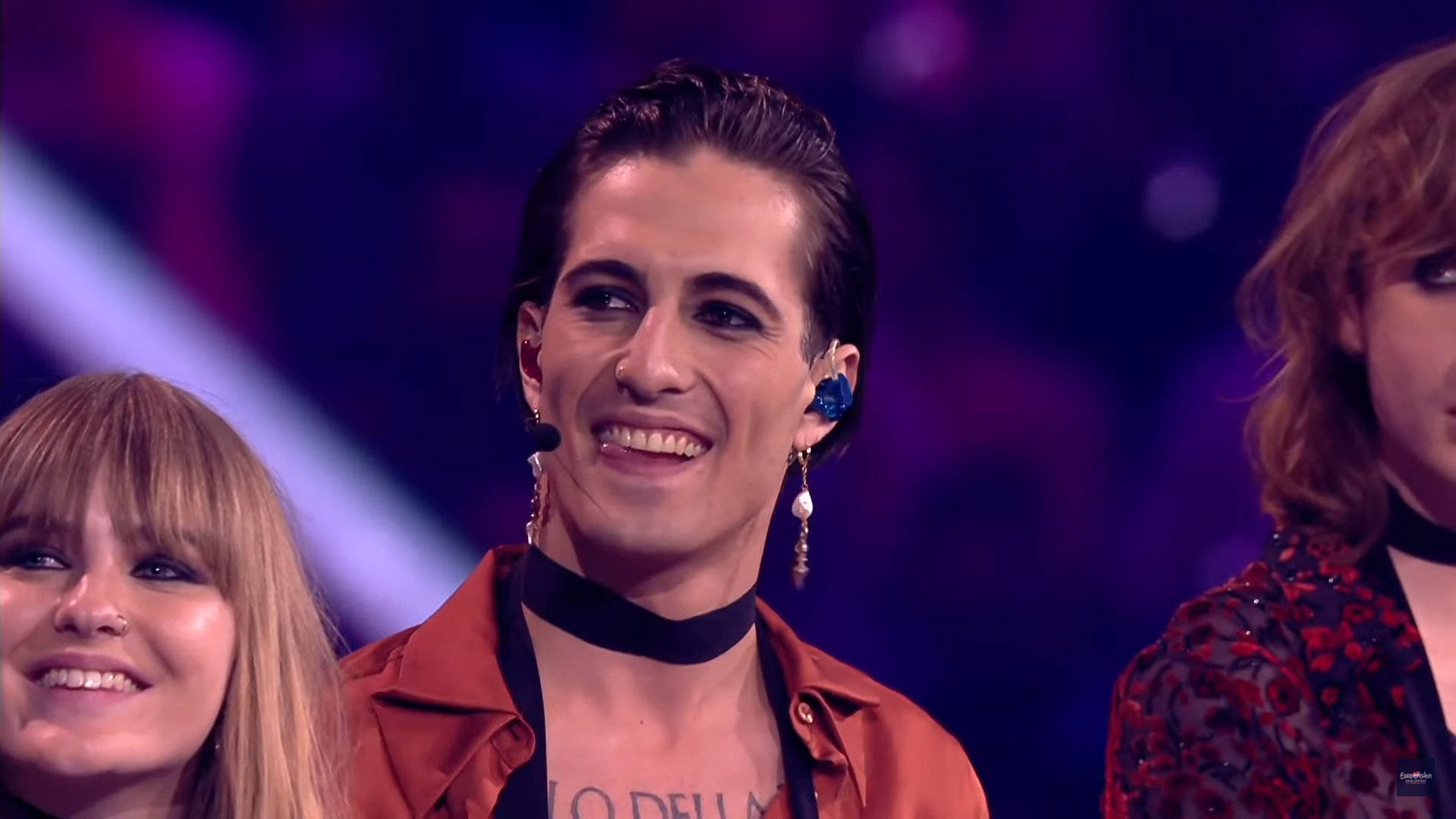 Maneskin 桌布 壁紙 — 一位面帶微笑的表演者，梳著時尚的髮型，戴著搖晃的耳環，在紫色舞台燈光下與另外兩位表演者一同閃耀。
