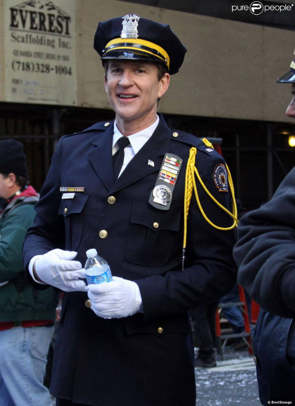 actor 桌布 壁紙 — 一位面帶微笑的警察，身穿深色制服，配有金色飾邊和白色手套，在城市的街道上拿著一個水瓶。