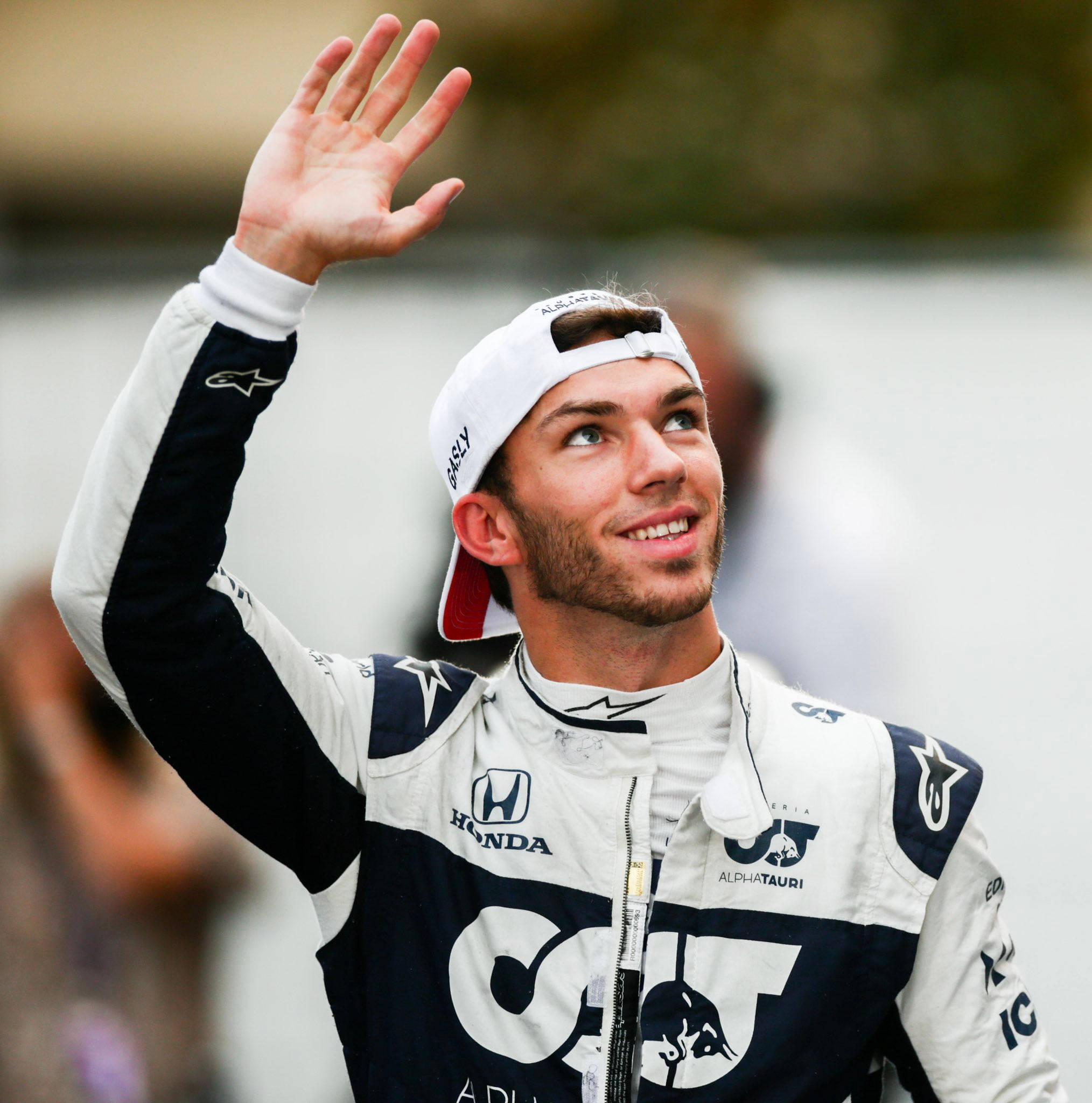 pierre gasly 桌布 壁紙 — 一位面帶微笑的賽車手身穿AlphaTauri Honda賽車服，戴著白色帽子向車迷揮手。