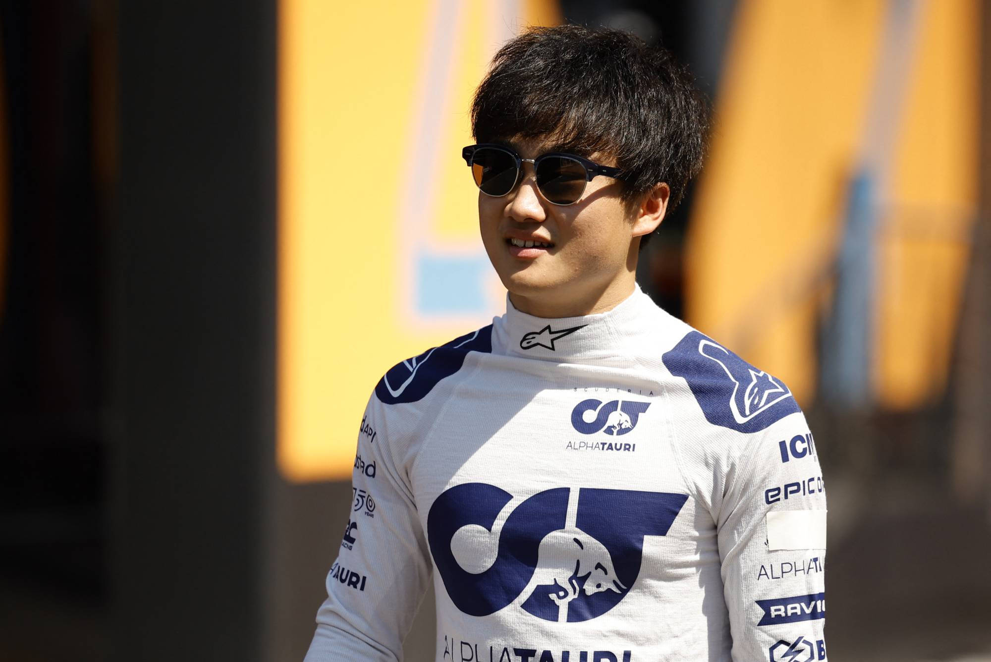 Sunglasses 桌布 壁紙 — 一位面帶微笑的賽車手身穿AlphaTauri車隊賽車服和太陽眼鏡，自信地站在模糊的黃色背景前。