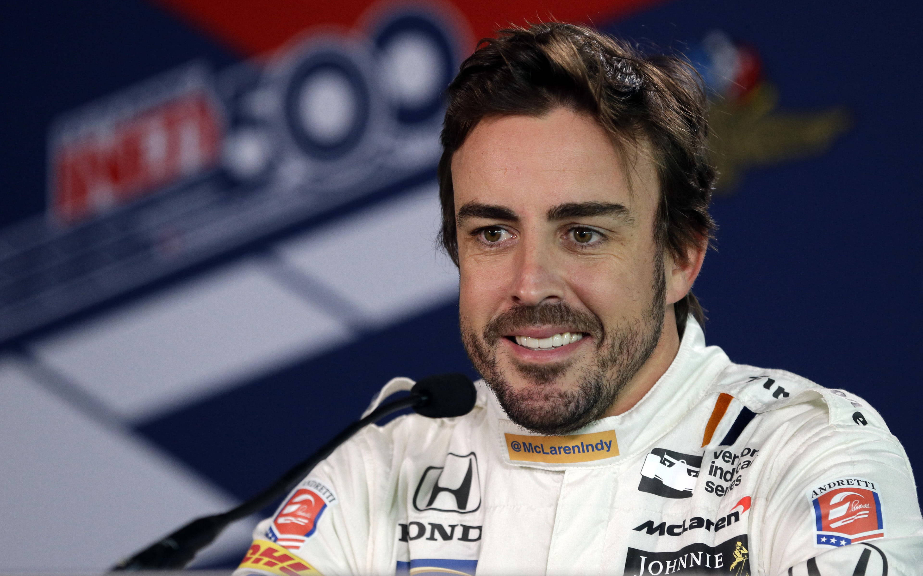 Fernando Alonso 桌布 壁紙 — 一位面帶微笑的賽車手身穿白色麥拉倫印地賽車制服，在記者會上發言，贊助商標誌清晰可見。
